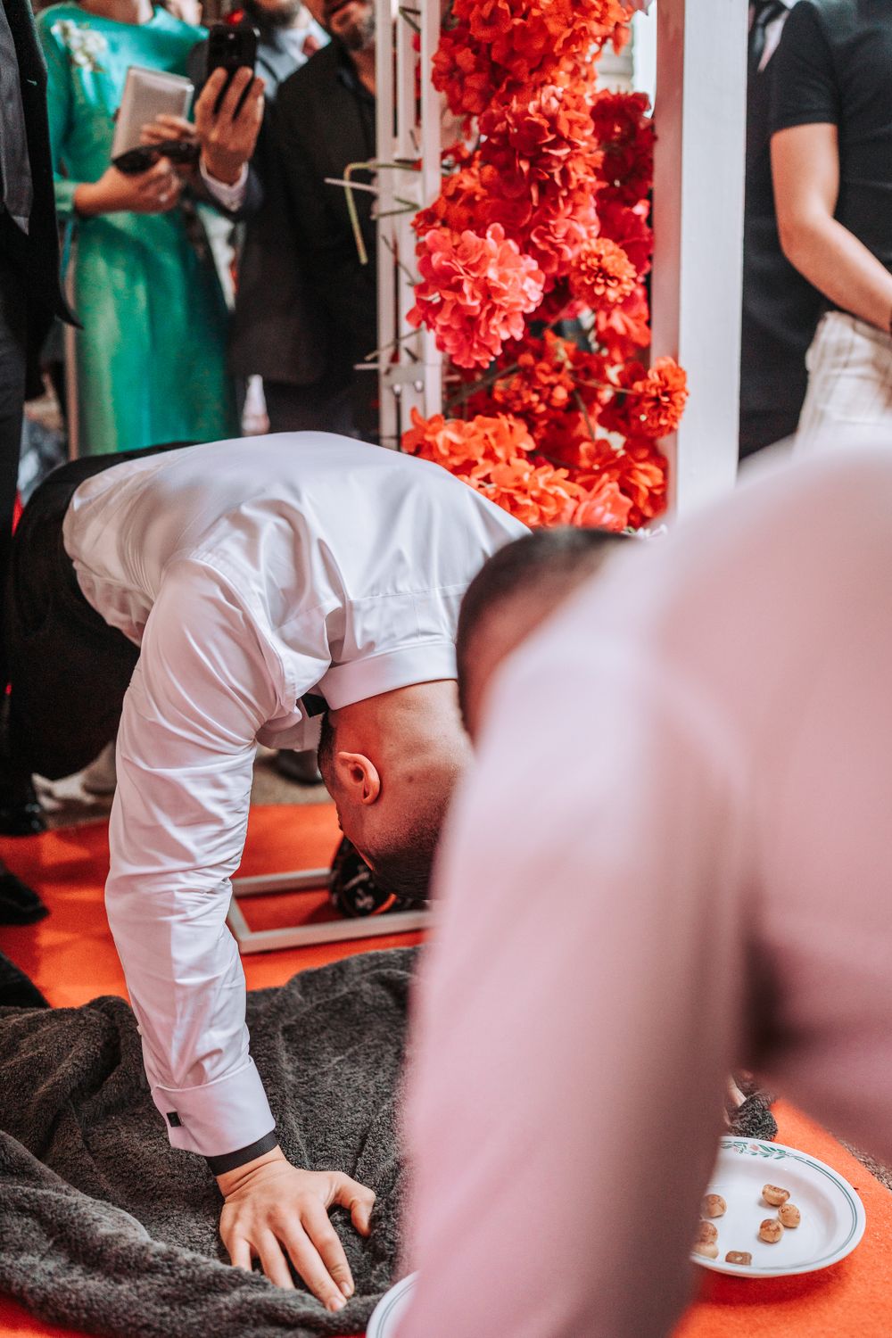 Person in white outfit bowing down during ceremony with red floral decorations in background.