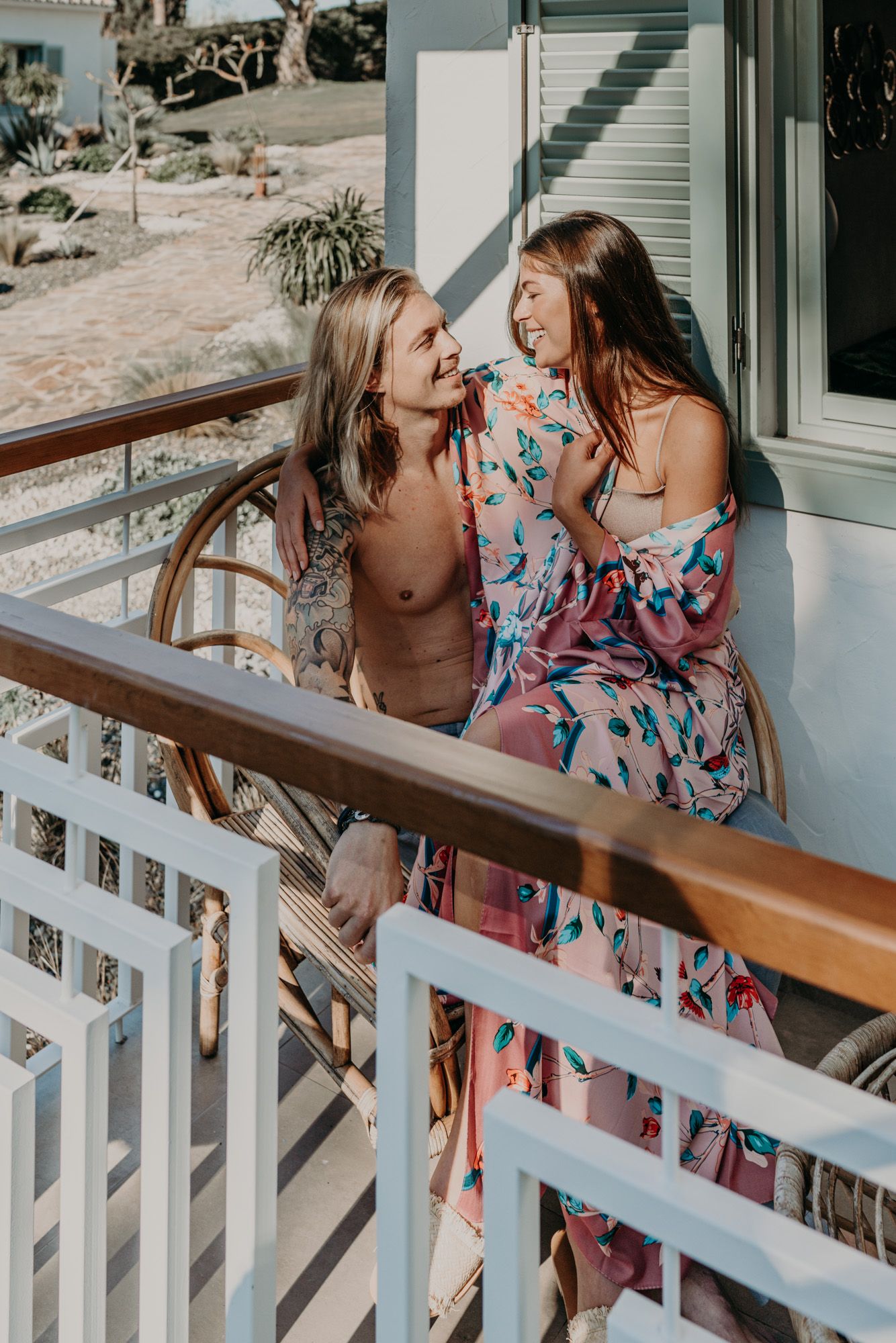 Two people share a lighthearted moment on a sunny porch, one wearing a floral dress and the other in casual attire.