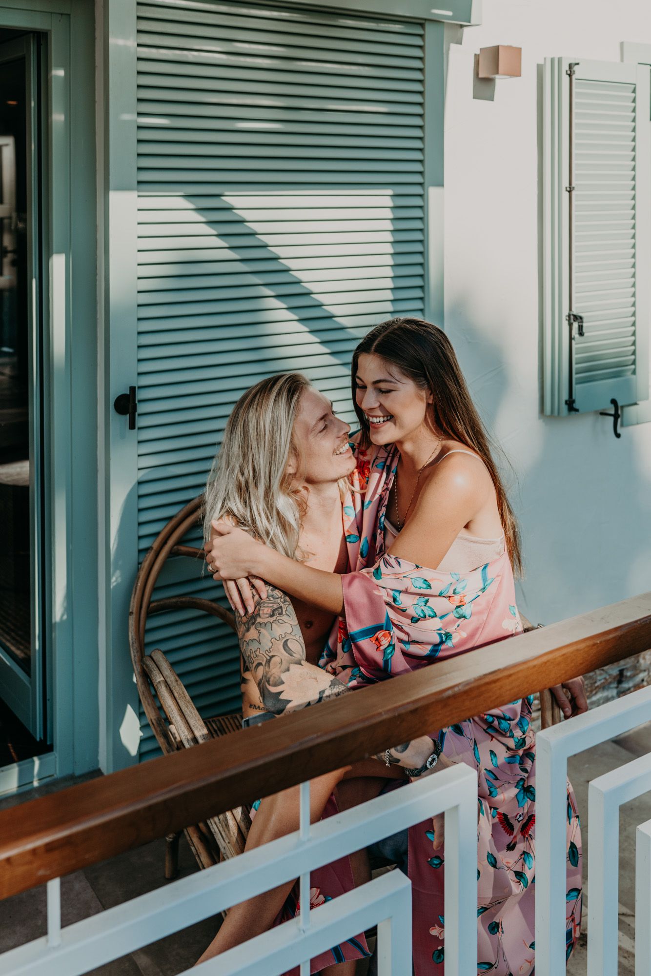 Two friends wearing summer dresses share a joyful moment on a balcony with teal shutters in the background.