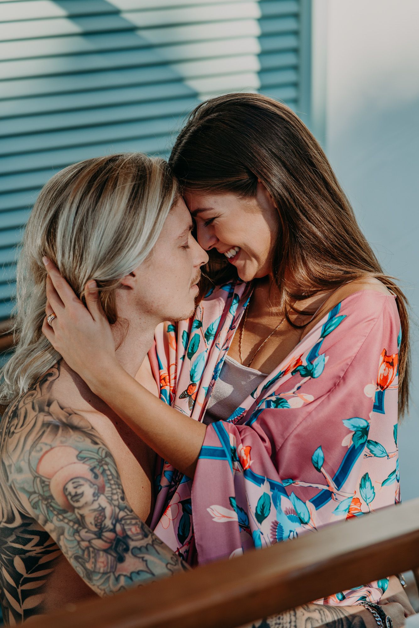 Two people sharing an intimate moment, one wearing a pink floral robe and the other with visible arm tattoos.