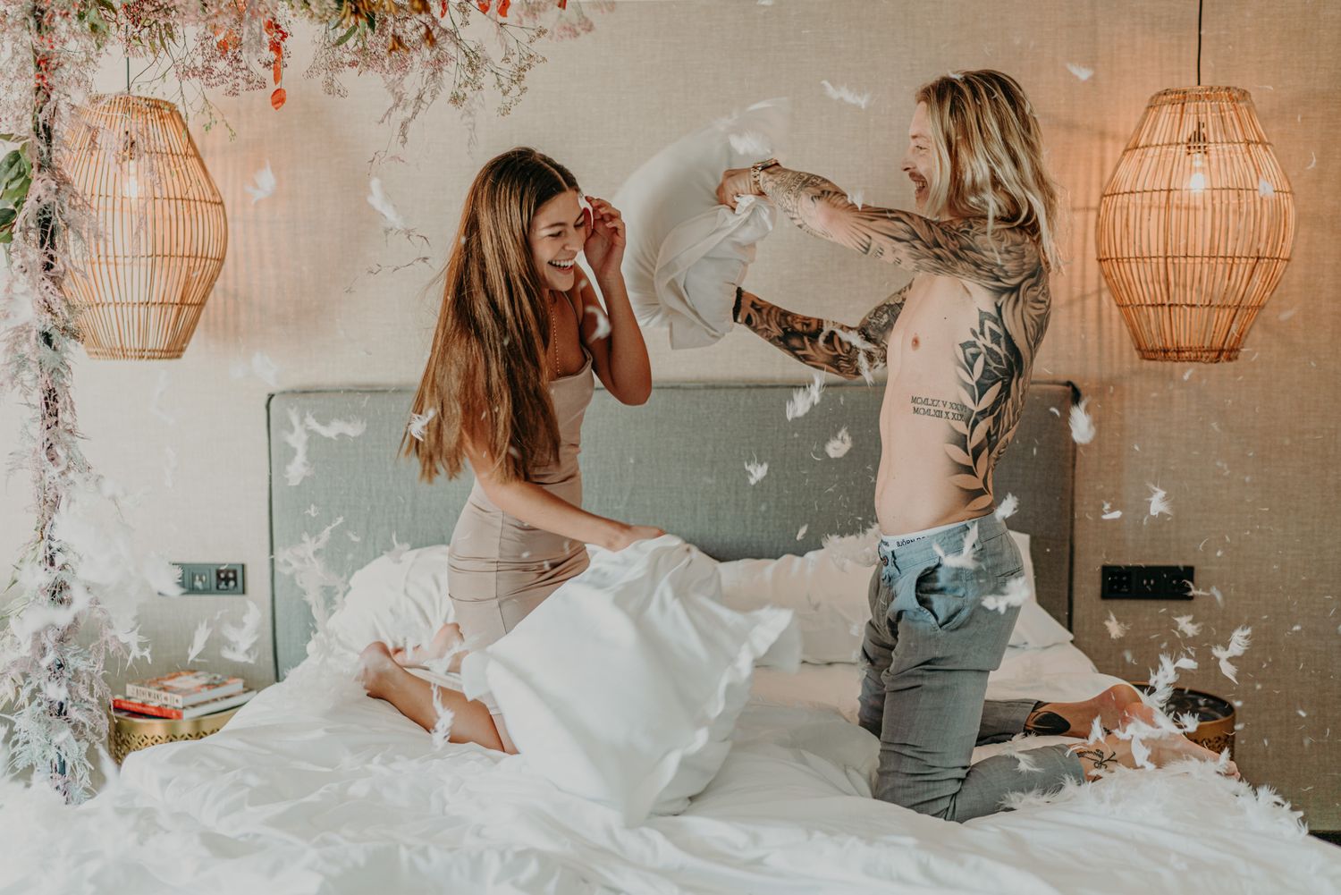 Two people having a playful pillow fight on a bed with decorative rattan lanterns hanging in the background.