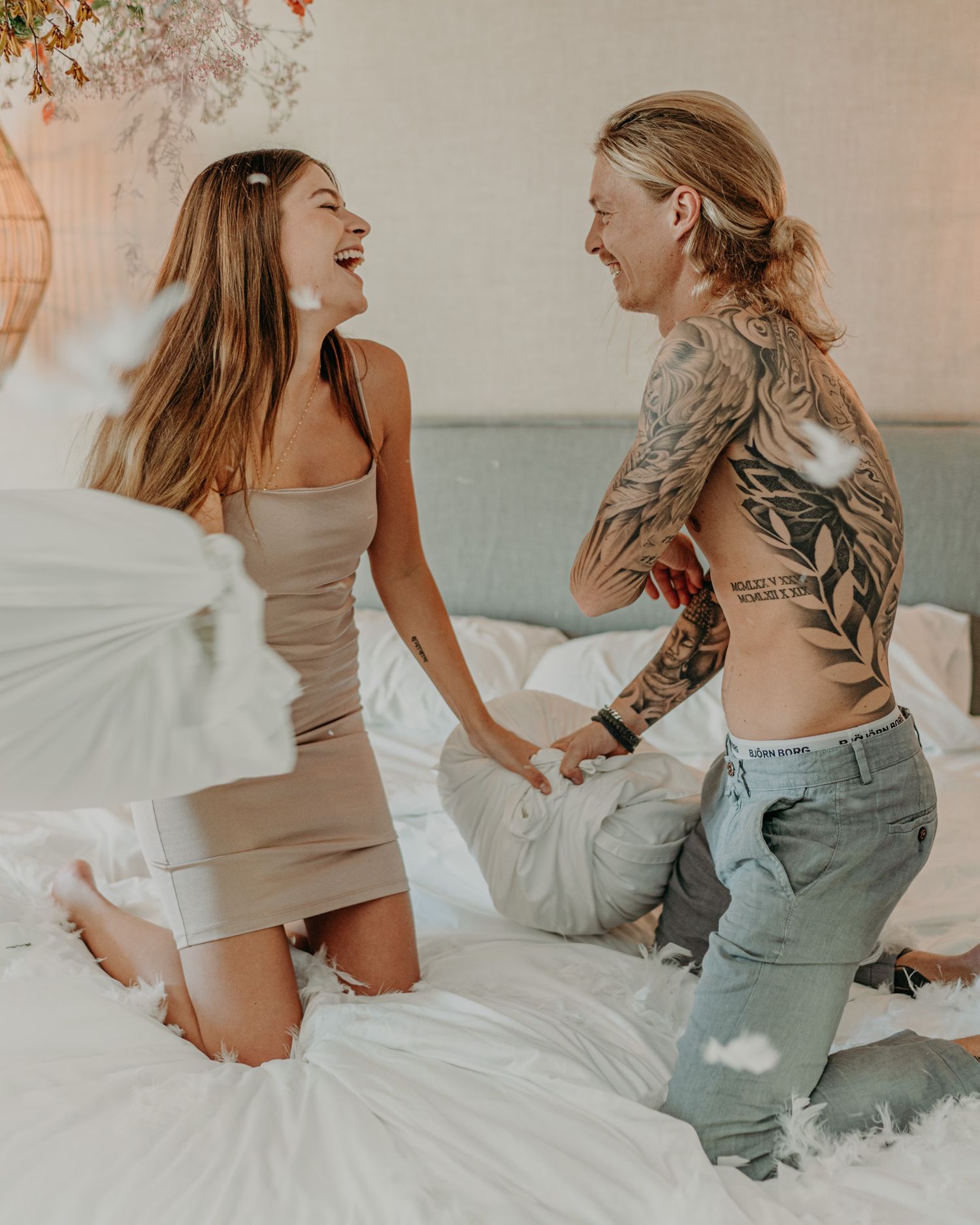 Two friends laughing and having fun during a playful pillow fight on a bed, one wearing a dress and one in denim.