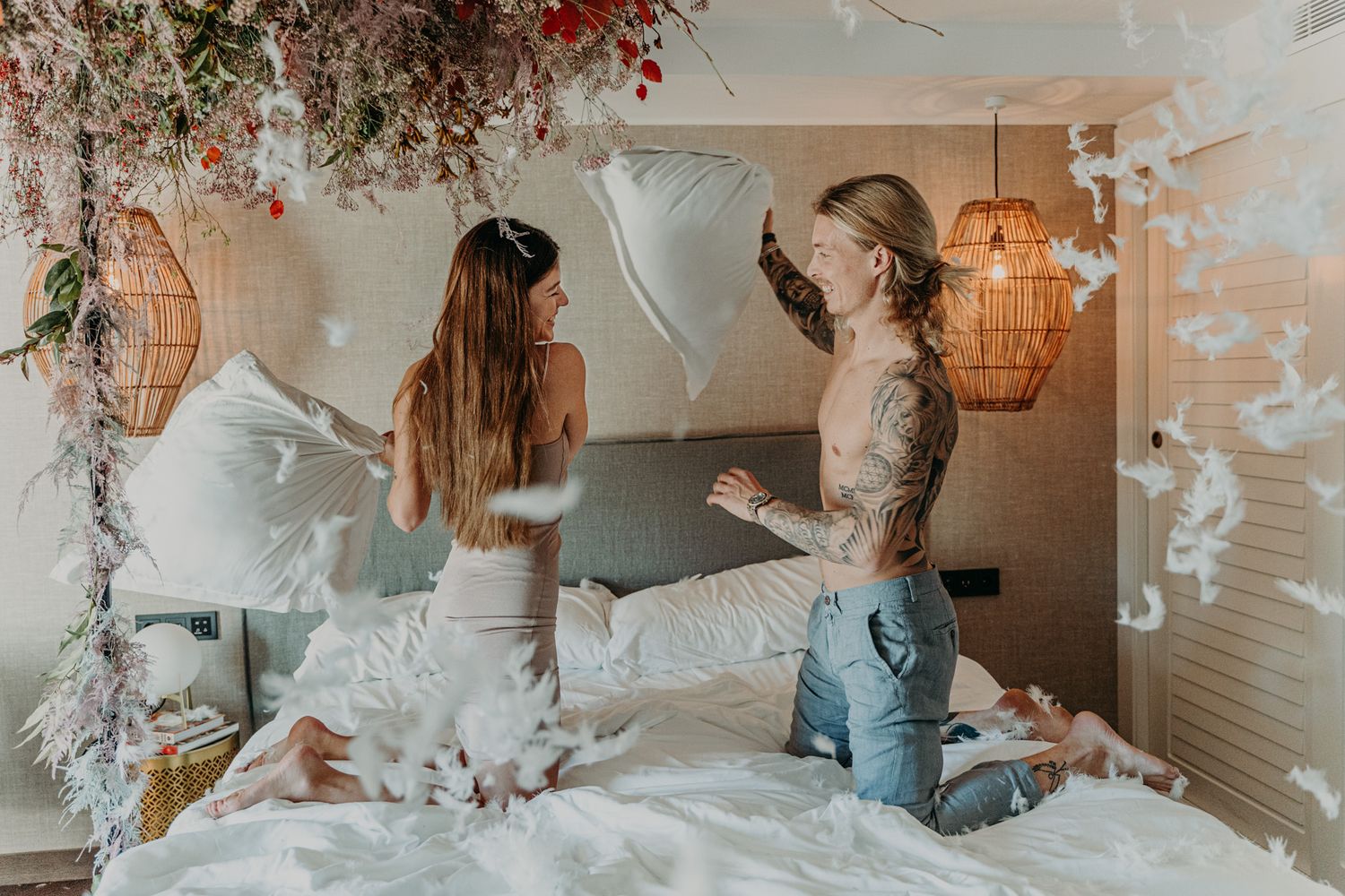 Two friends having a playful pillow fight on a bed in a cozy bedroom decorated with rattan lamps and floral accents.