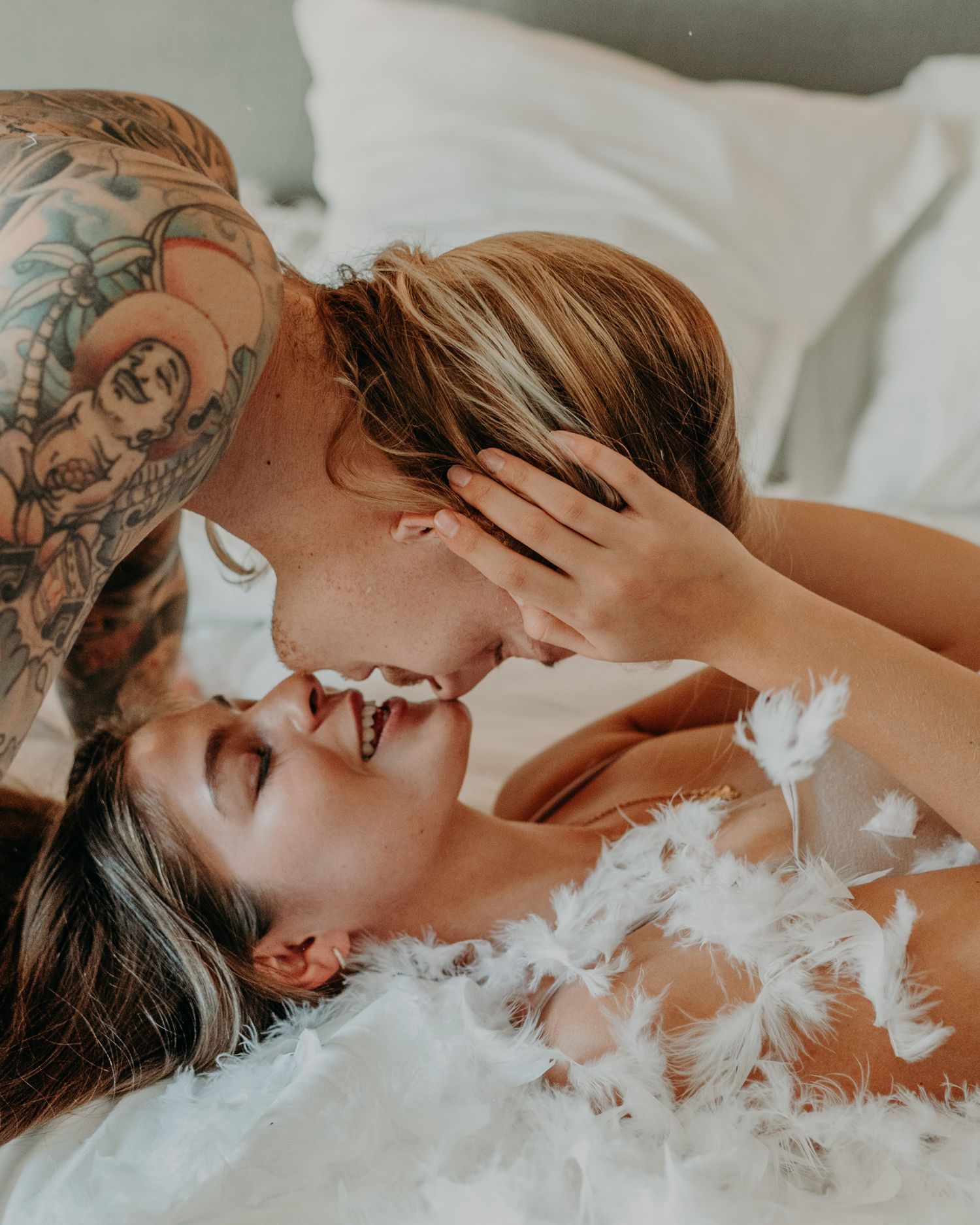 A romantic moment captured on white bedding with feathers and visible tattoo artwork in warm lighting.