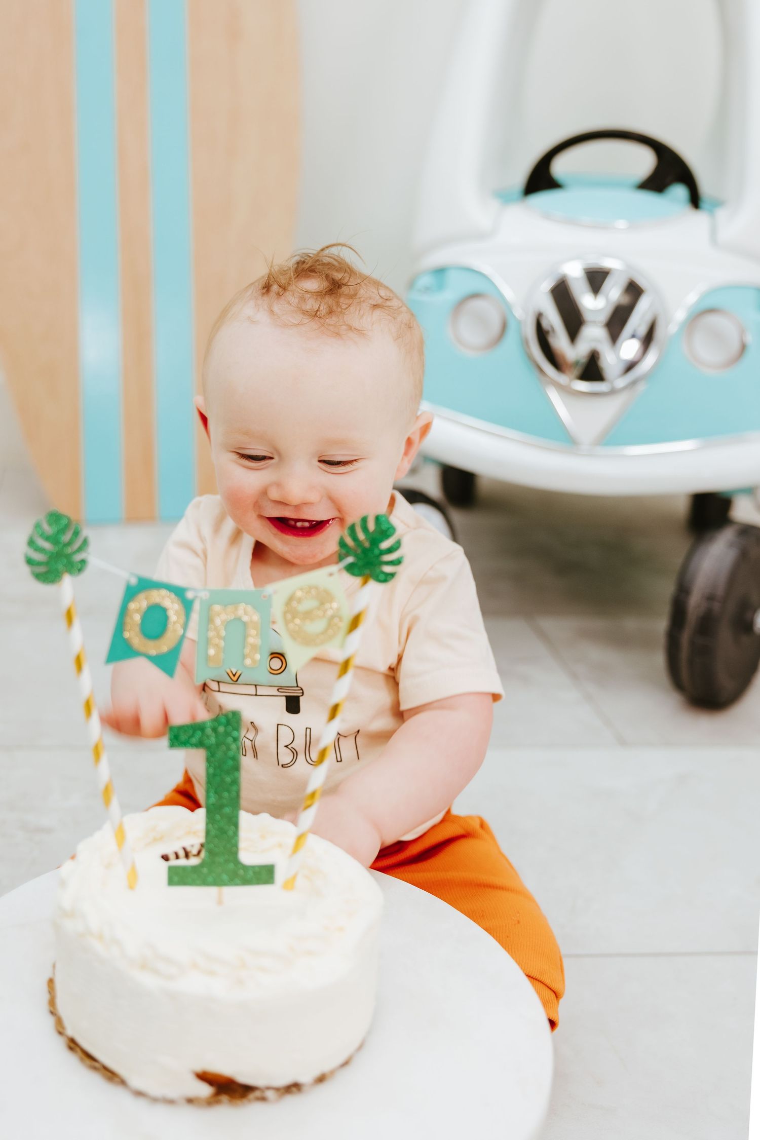 A first birthday celebration with white cake and decorative number one topper next to a light blue toy VW bus.