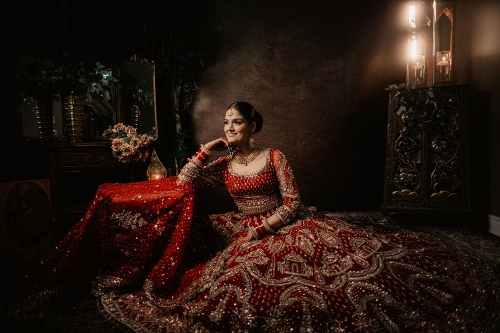 A person wearing an ornate red and gold Indian bridal lehenga sits elegantly in a dimly lit vintage-style room.