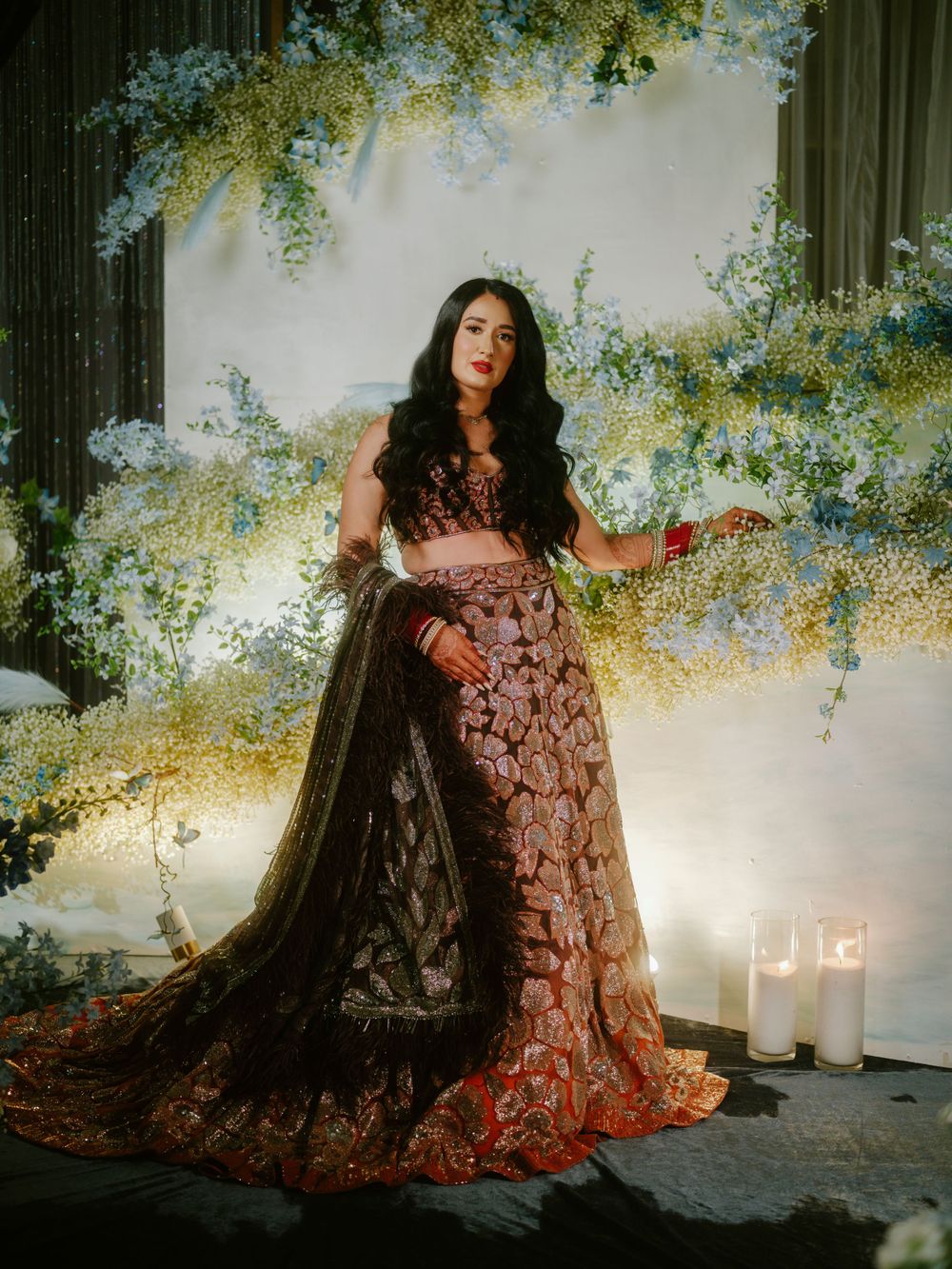 Ethereal fashion photograph in a romantic garden setting with flowing embroidered gown and dramatic black cape amid candles.