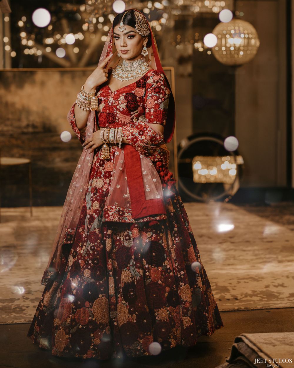 Traditional red and gold bridal lehenga with floral embellishments and sparkling decor in dimly lit ballroom setting.