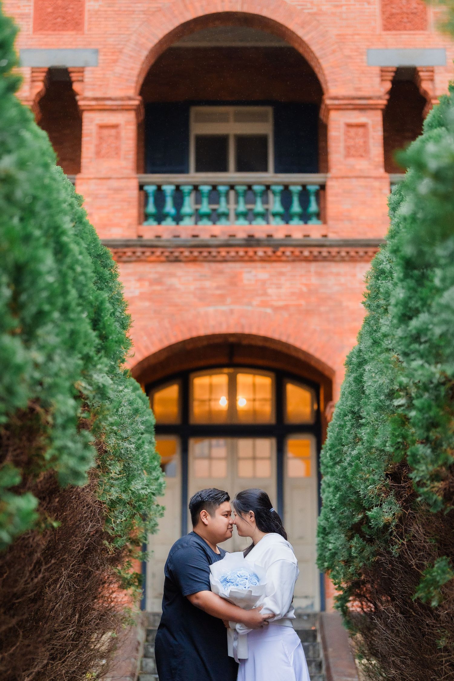A couple embraces outside a historic brick building with arched windows and tall cypress trees framing the entrance.