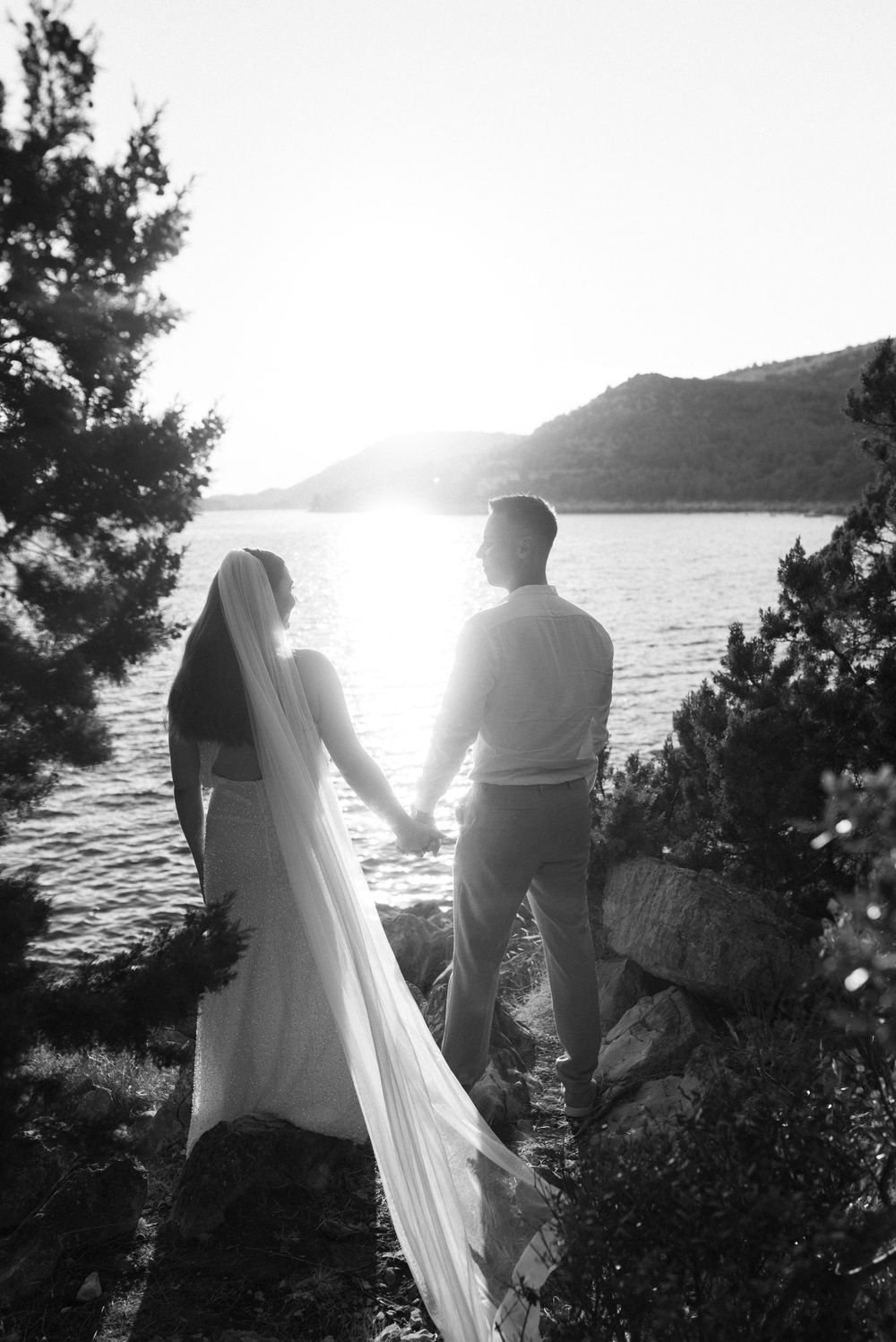 Black and white silhouette of couple holding hands while overlooking sunset on rocky shore.