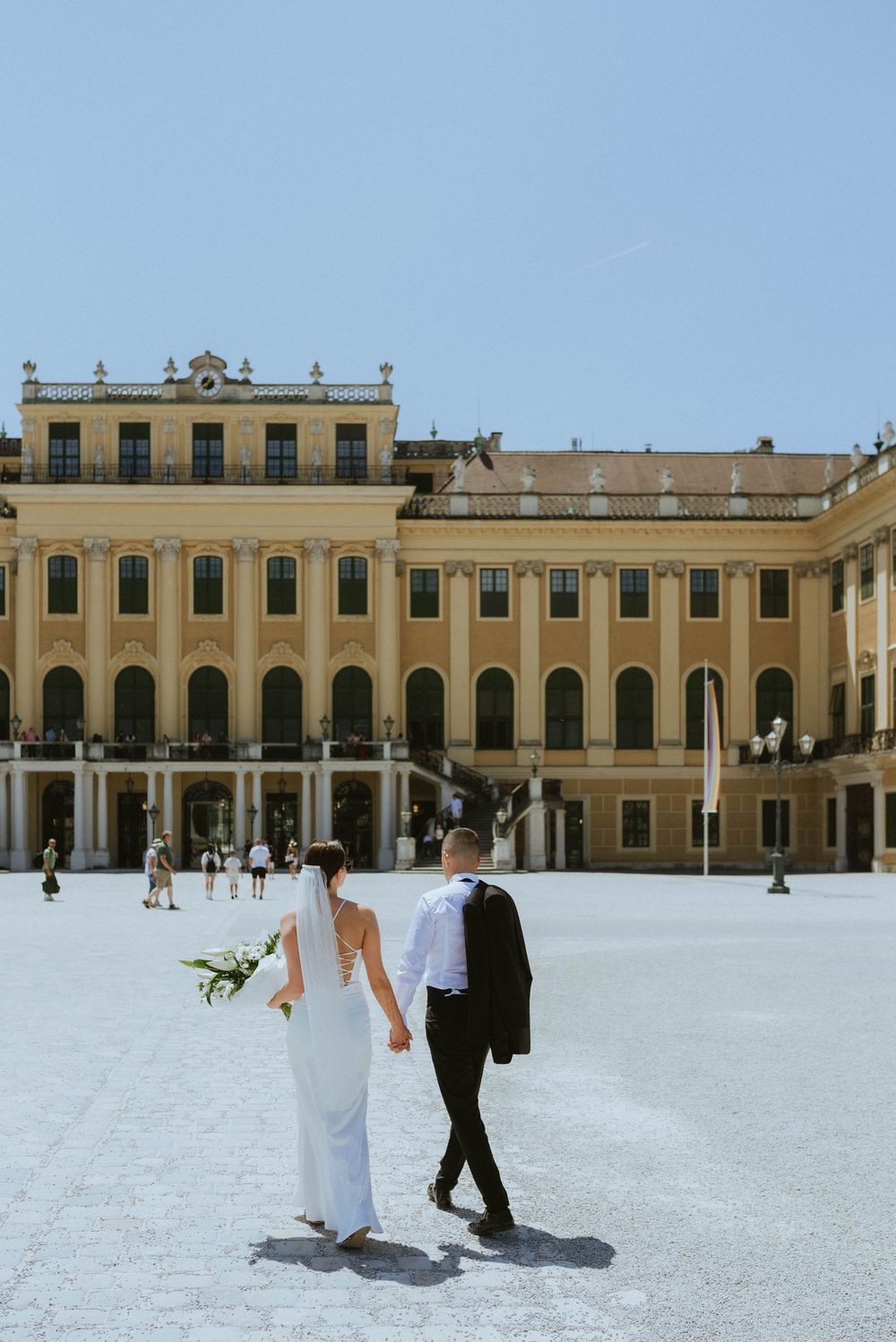 A couple walks in elegant attire across the courtyard of Schönbrunn Palace in Vienna on a sunny day.