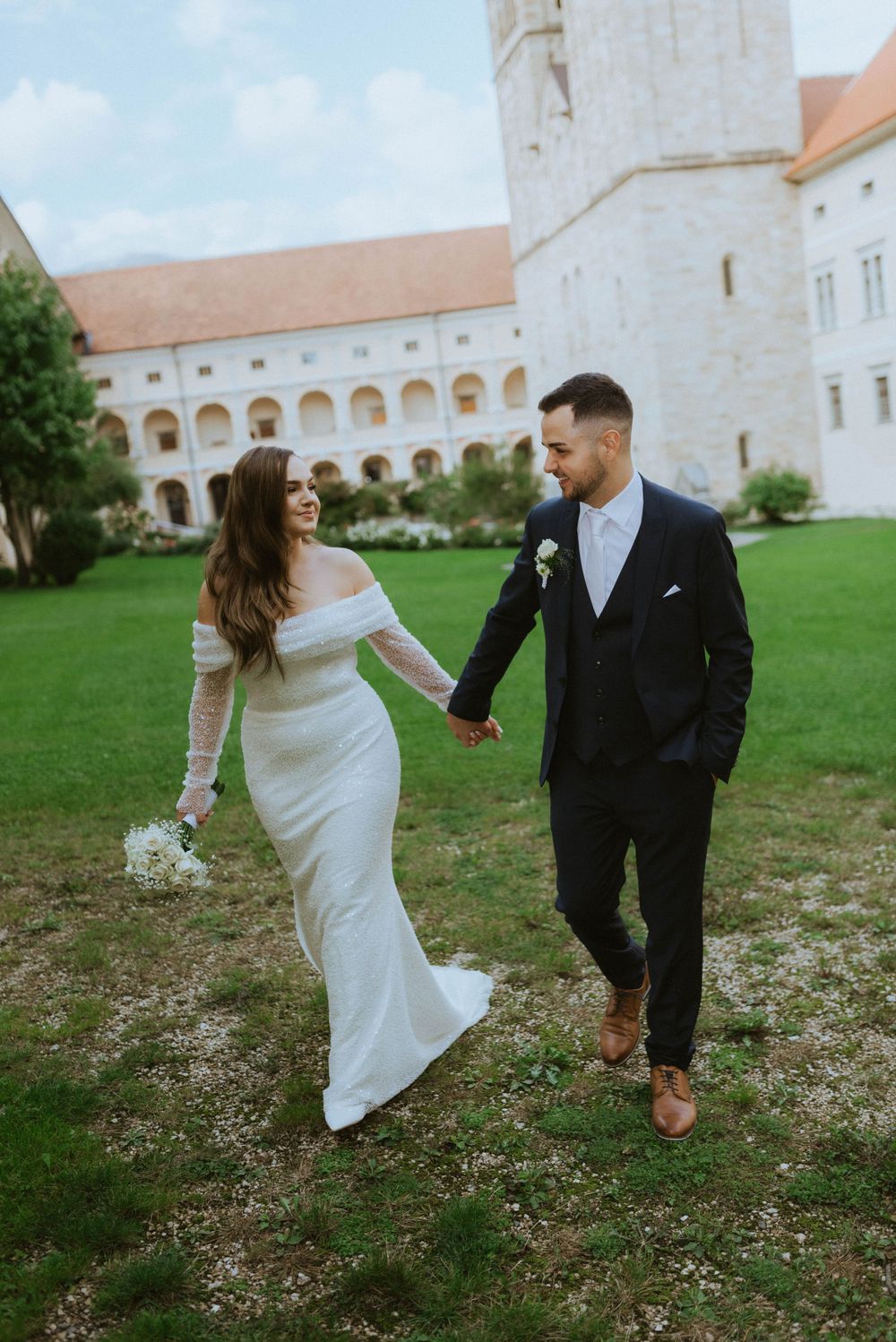 A wedding couple walks hand in hand on a grassy lawn in front of a historic monastery with white walls and arches.