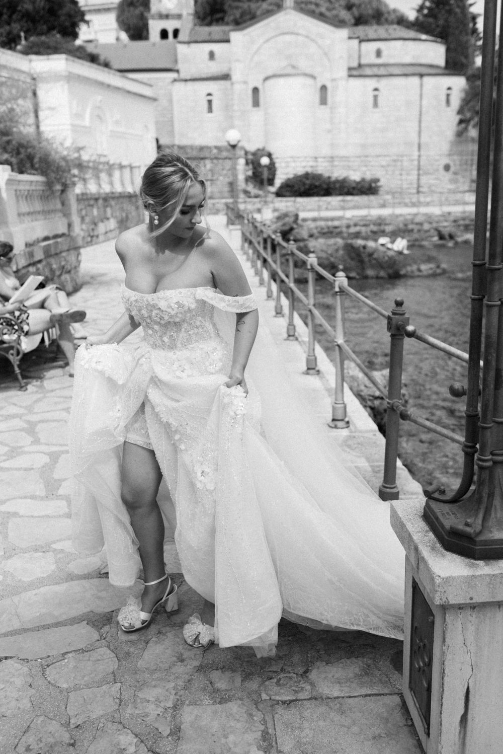 Bride in lace dress lifting her gown while standing on stone steps near historic building.