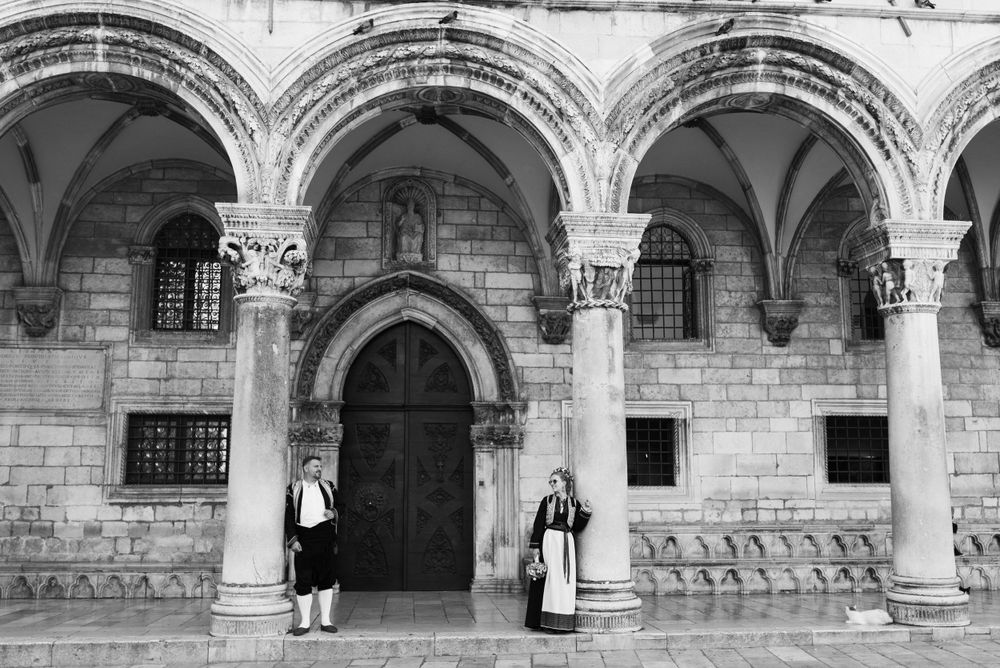 A black and white photo showing traditional dress at a historic stone archway entrance.