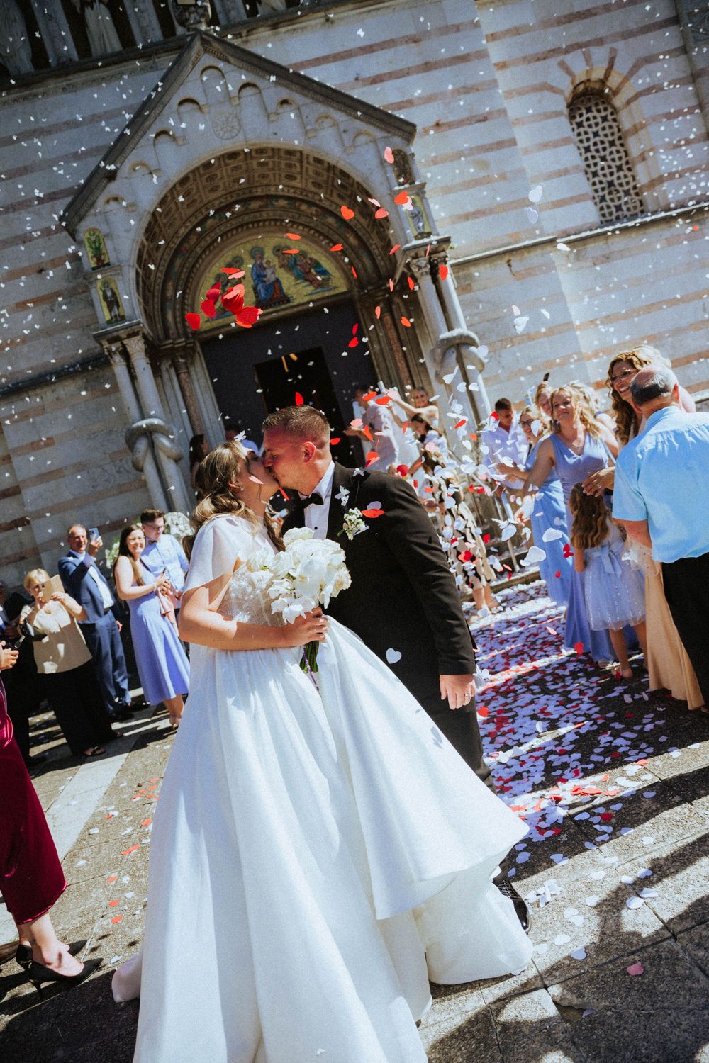 Newlyweds share a kiss outside a church as guests throw colorful confetti, bride holding white bouquet in elegant gown.