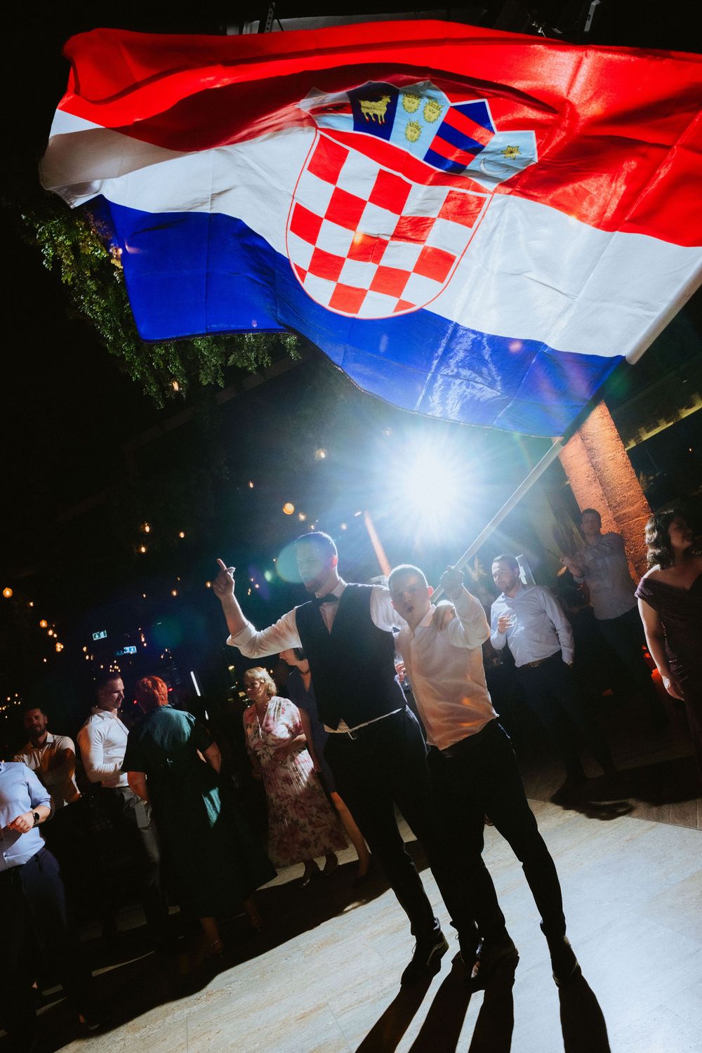 People dancing at a night event under a waving Croatian flag with red and white checkerboard coat of arms.