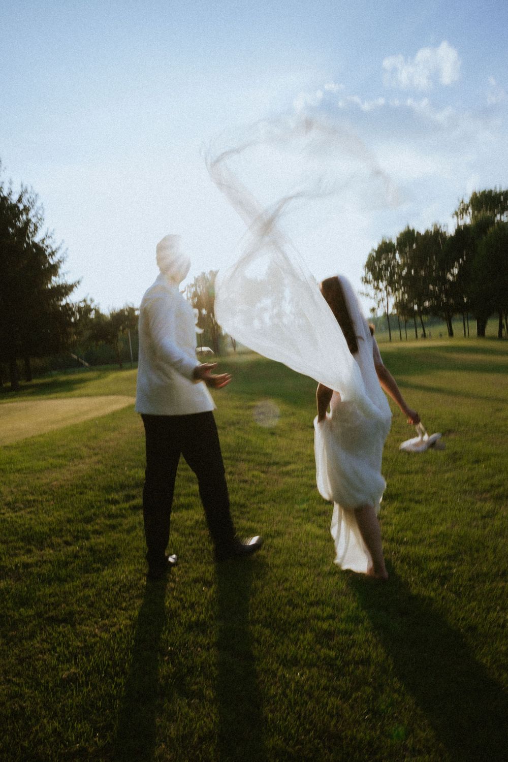 White flowing wedding dress catches wind on golf course at sunset creating ethereal silhouette against blue sky.