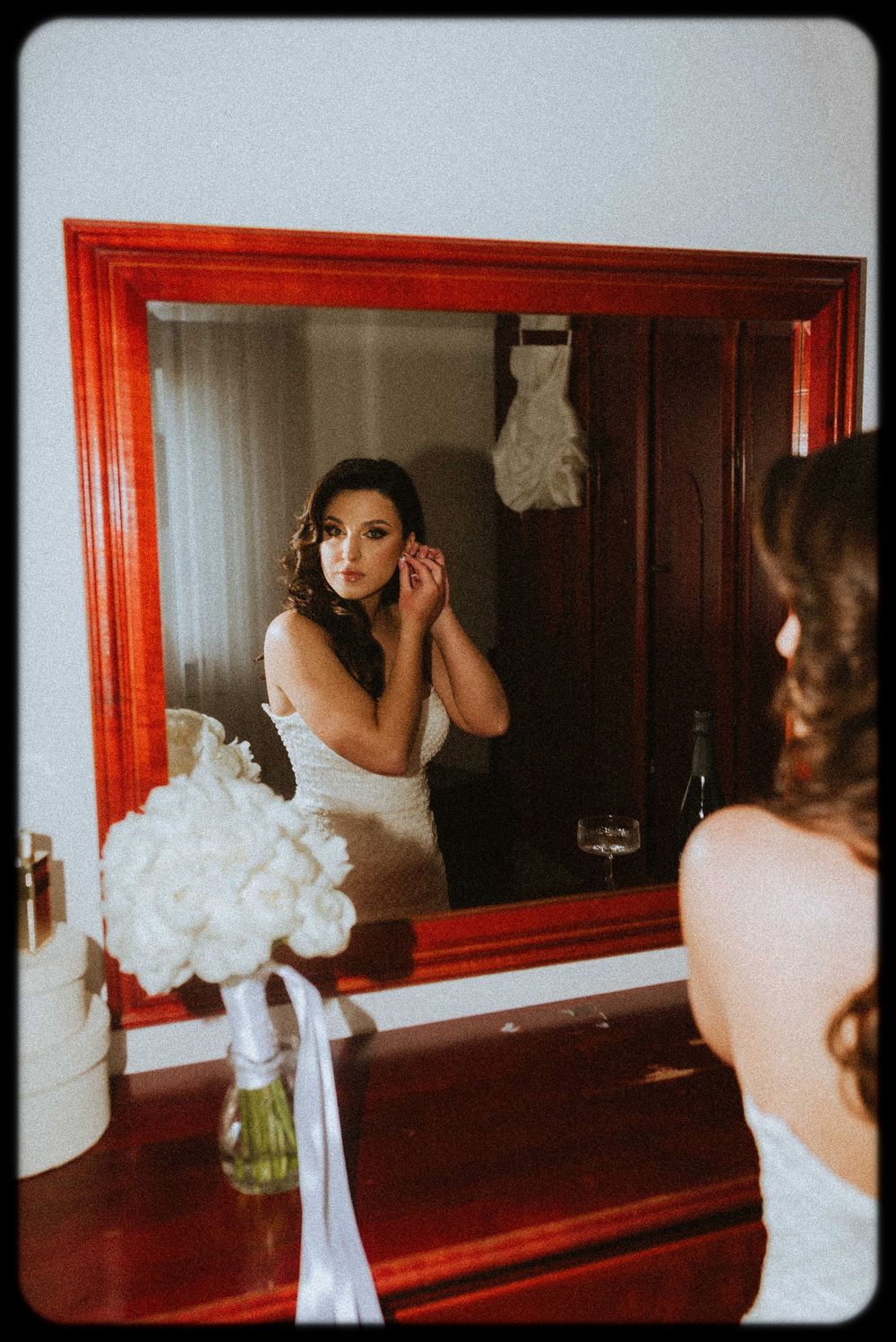 A bride in a white wedding dress puts on earrings while looking in a red-framed mirror with a white bouquet visible.