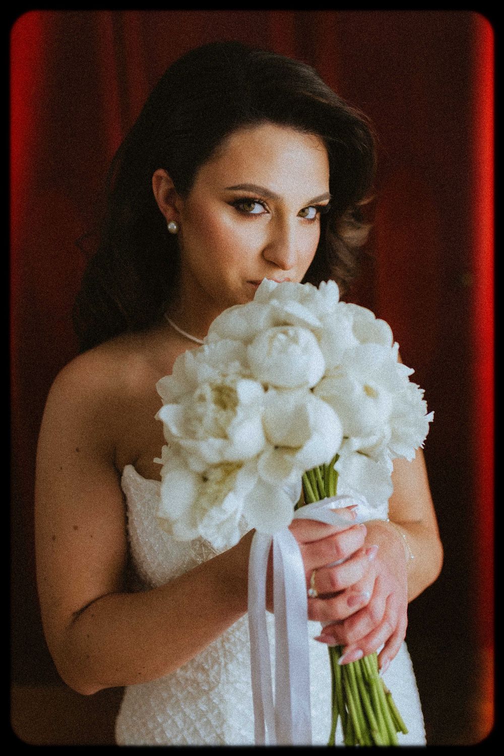 A bride in a white lace dress holds a classic bouquet of white peonies against a red backdrop.