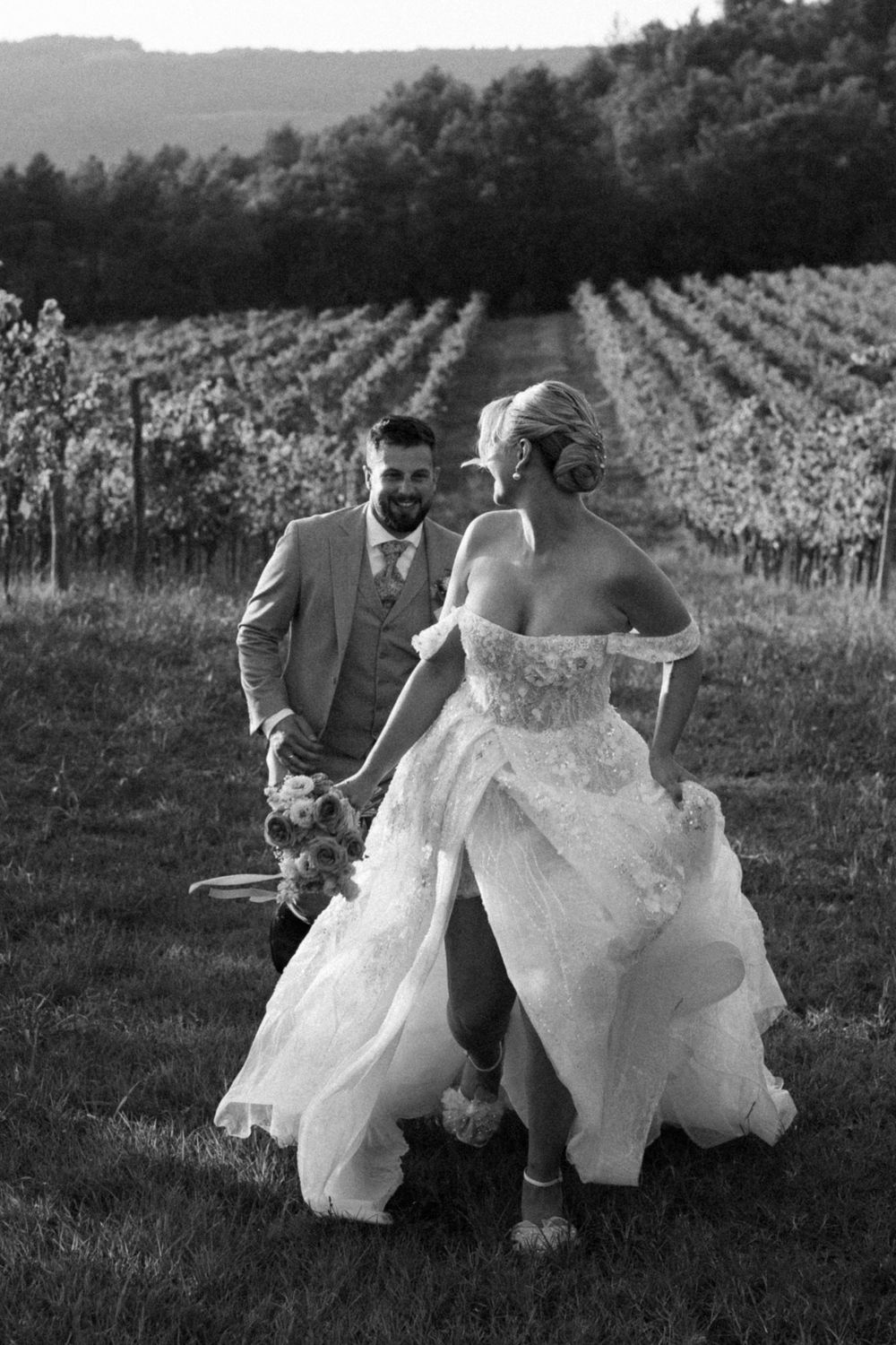 Wedding couple running playfully through vineyard rows at sunset.