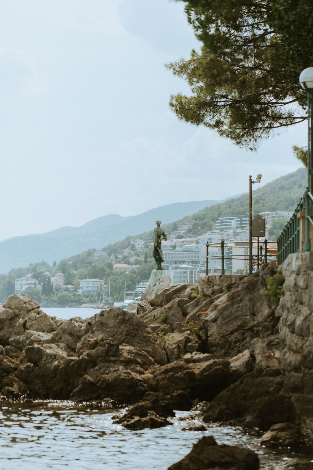 A bronze statue stands on rocky coastal cliffs overlooking a Mediterranean seaside town.