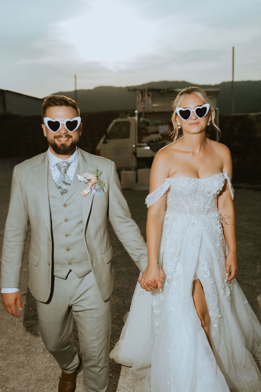 A couple wearing sunglasses and wedding attire poses together at sunset.