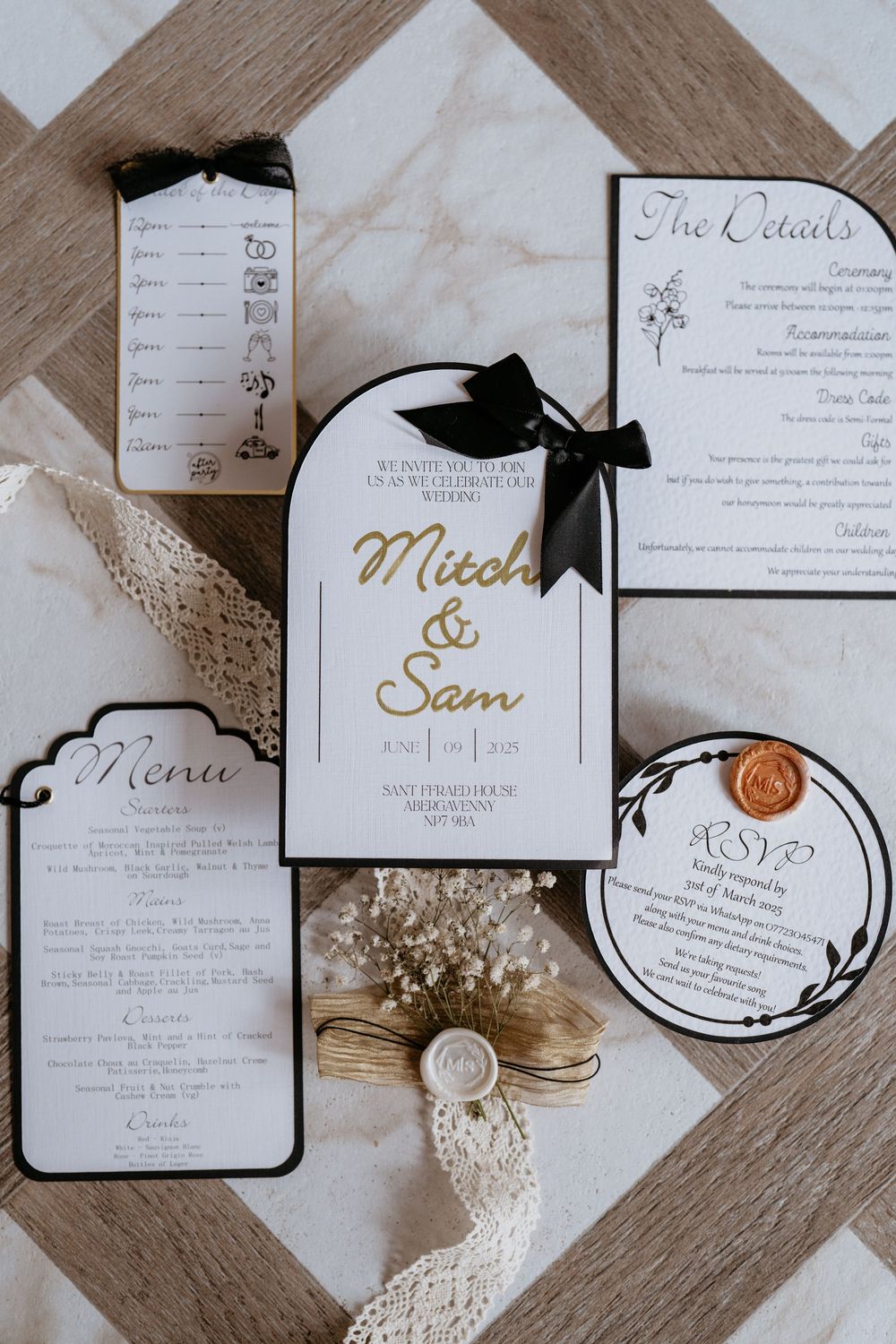 Elegant wedding stationery suite featuring black and gold details with wax seals and lace accents laid out on wooden surface. AT SANT FFRAED