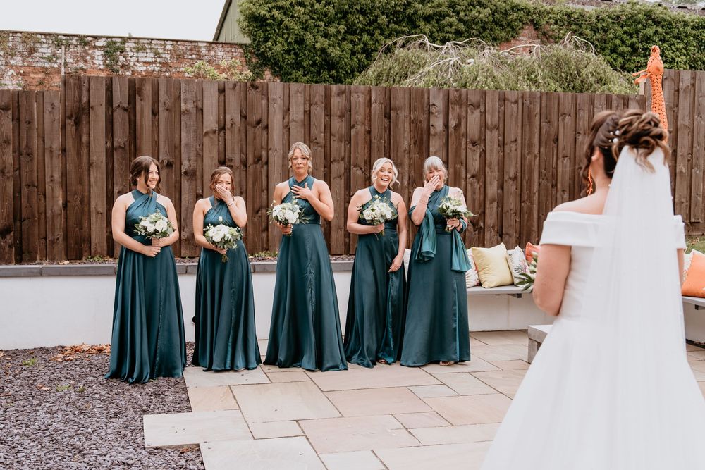 A bride in a white wedding gown faces bridesmaids wearing emerald green dresses in an outdoor courtyard setting. AT SANT FFRAED HOUSE