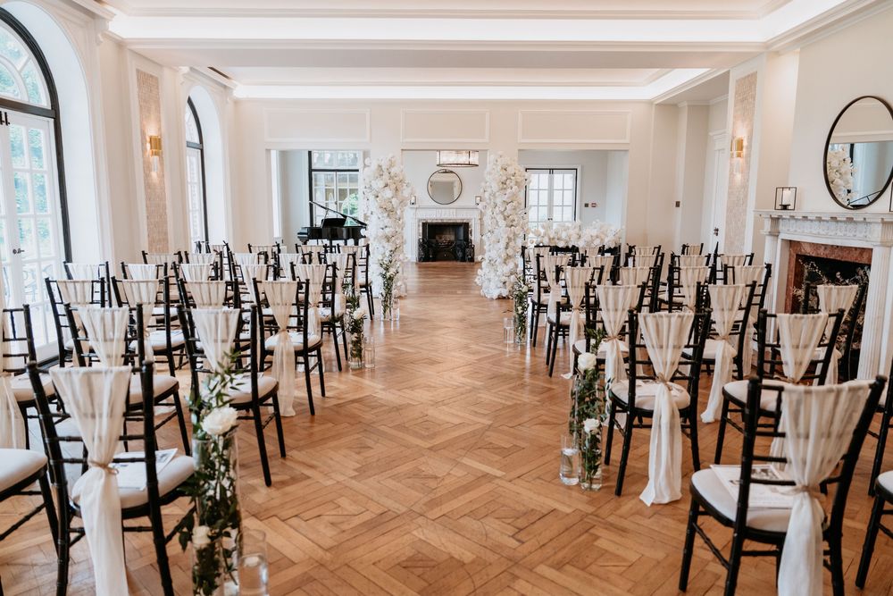 Panoramic view of an elegant ballroom with herringbone wood floors and black Chiavari chairs arranged in rows. AT SANT FFRAED HOUSE