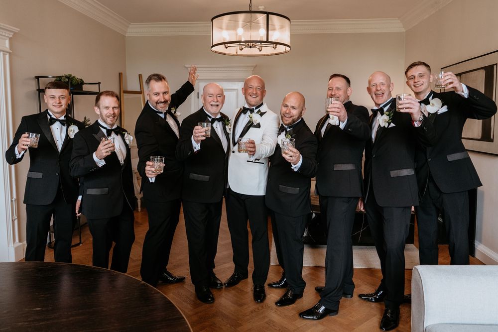 Panoramic group photo of wedding party members in formal black attire posing with playful gestures along a white wall. AT SANT FFRAED HOUSE