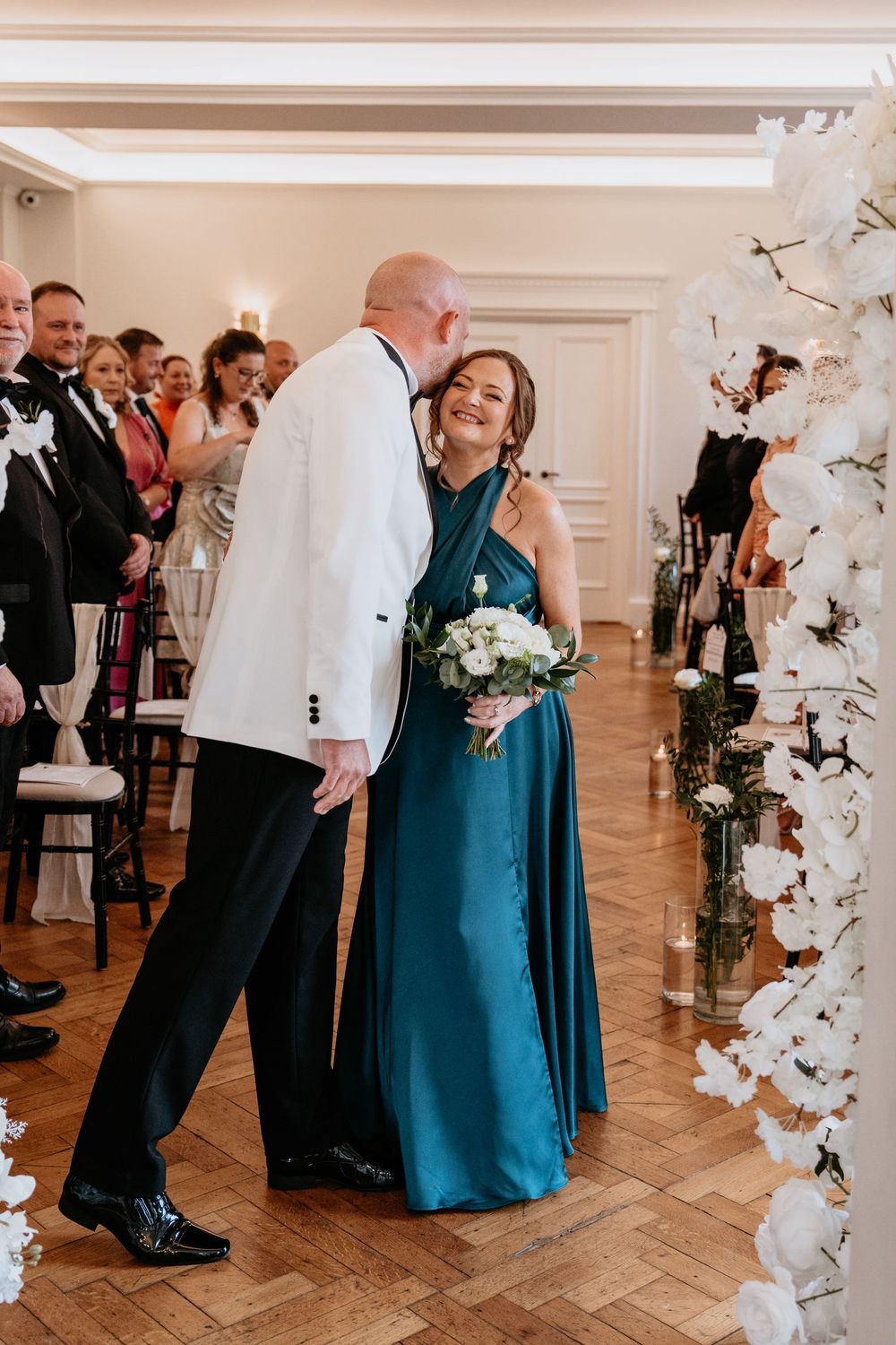 A couple shares an intimate moment during their wedding reception against a backdrop of white floral decorations. AT SANT FFRAED HOUSE