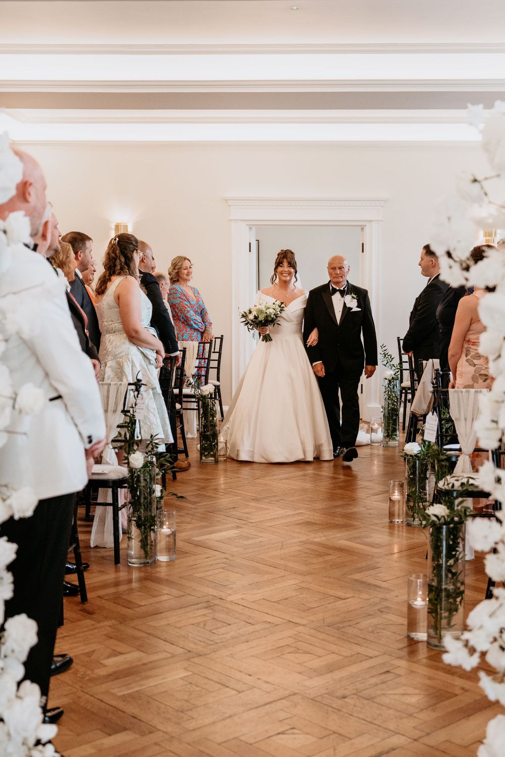 Wedding couple walking down aisle in elegant indoor venue with parquet flooring and floral arrangements. AT SANT FFRAED HOUSE