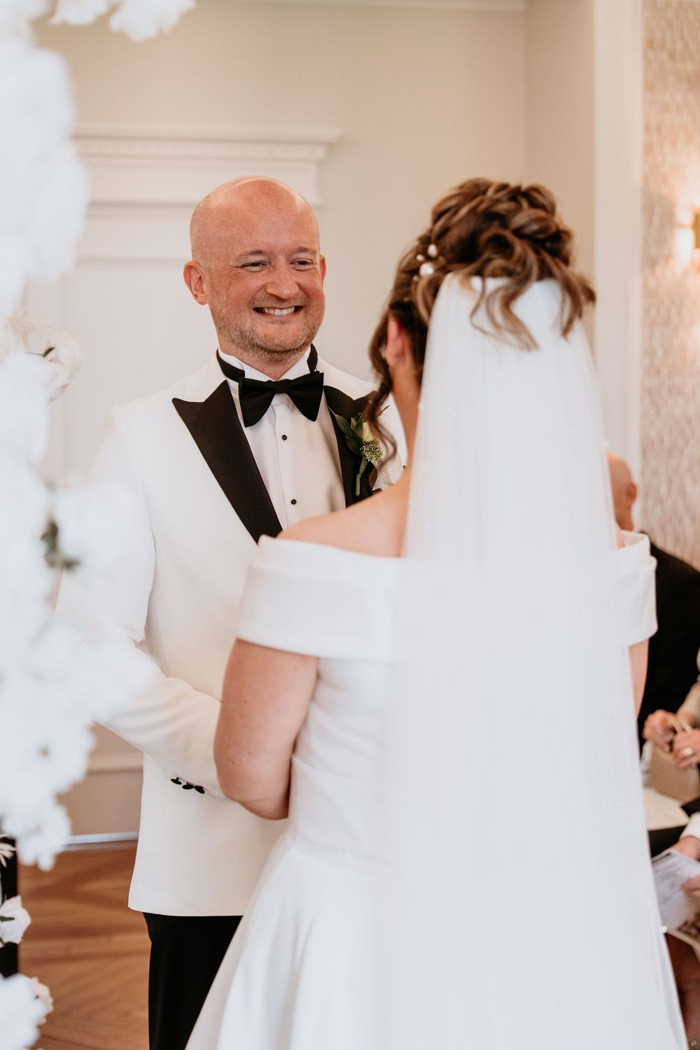 At a wedding ceremony, a couple shares an intimate moment in formal attire against an elegant white backdrop with floral decor. SANT FFRAED
