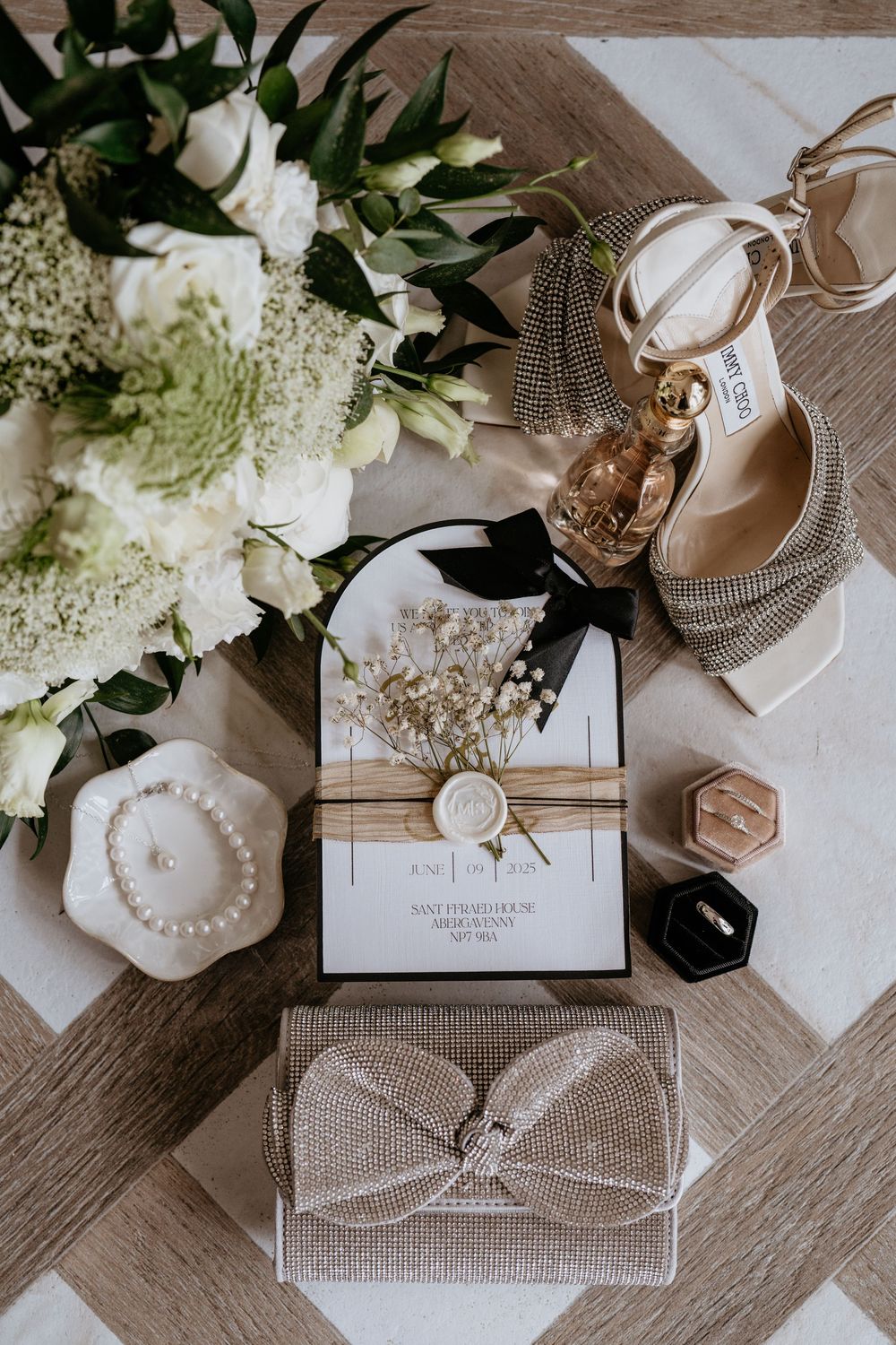 Elegant wedding details including sparkly heels, white floral bouquet, invitation suite and decorative boxes on wooden surface.SANT FFRAED