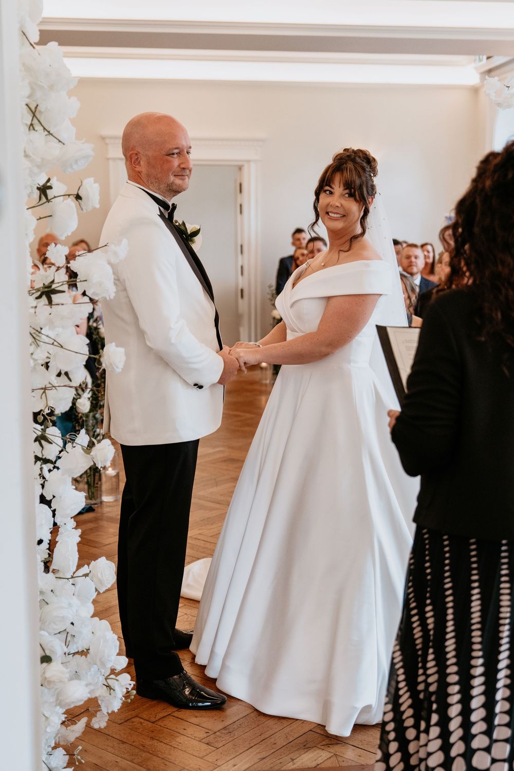 A wedding ceremony in a bright indoor venue decorated with white floral arrangements. AT SANT FFRAED HOUSE