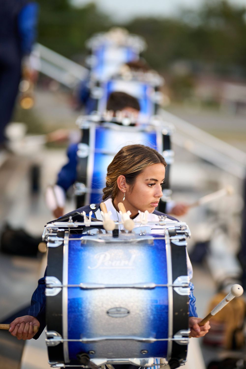 Drummer in blue marching band uniform performs with large bass drum during outdoor event.