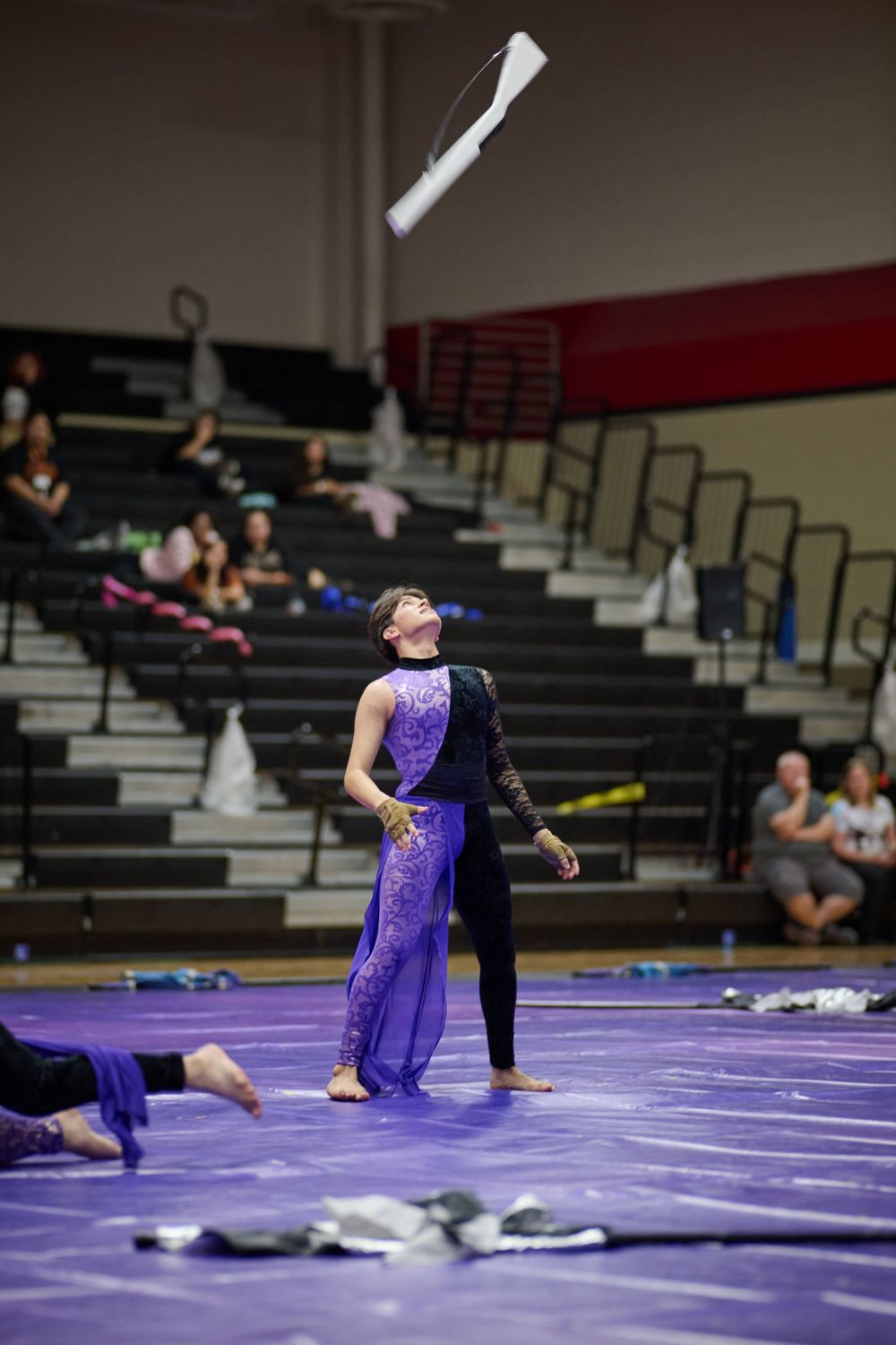 Color guard performer in purple costume tossing equipment during indoor winter guard show.