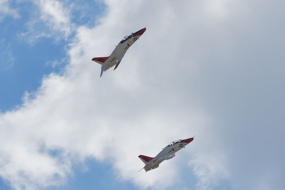 Two military jets performing an aerial maneuver against a cloudy blue sky.