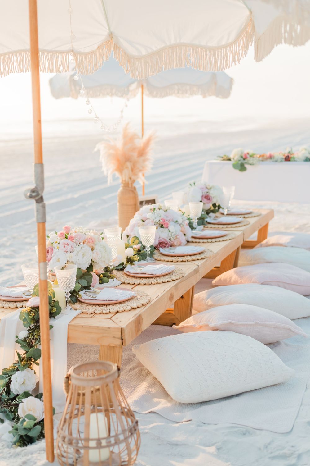 Romantic beach picnic setup with white cushions, wooden table, floral decor and umbrella on sand during sunset.