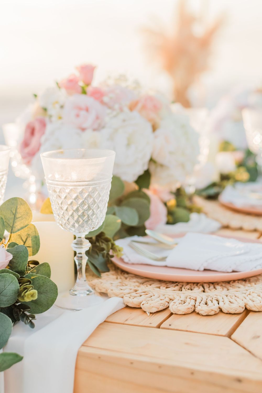 Elegant table setting with crystal goblet, pink roses, eucalyptus leaves and lace doilies on wooden surface.