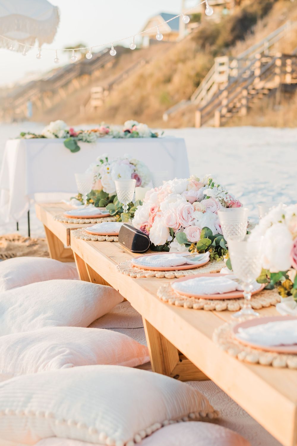 Elegant beach dining setup with wooden table, plush floor cushions, and delicate pink floral centerpieces at sunset.
