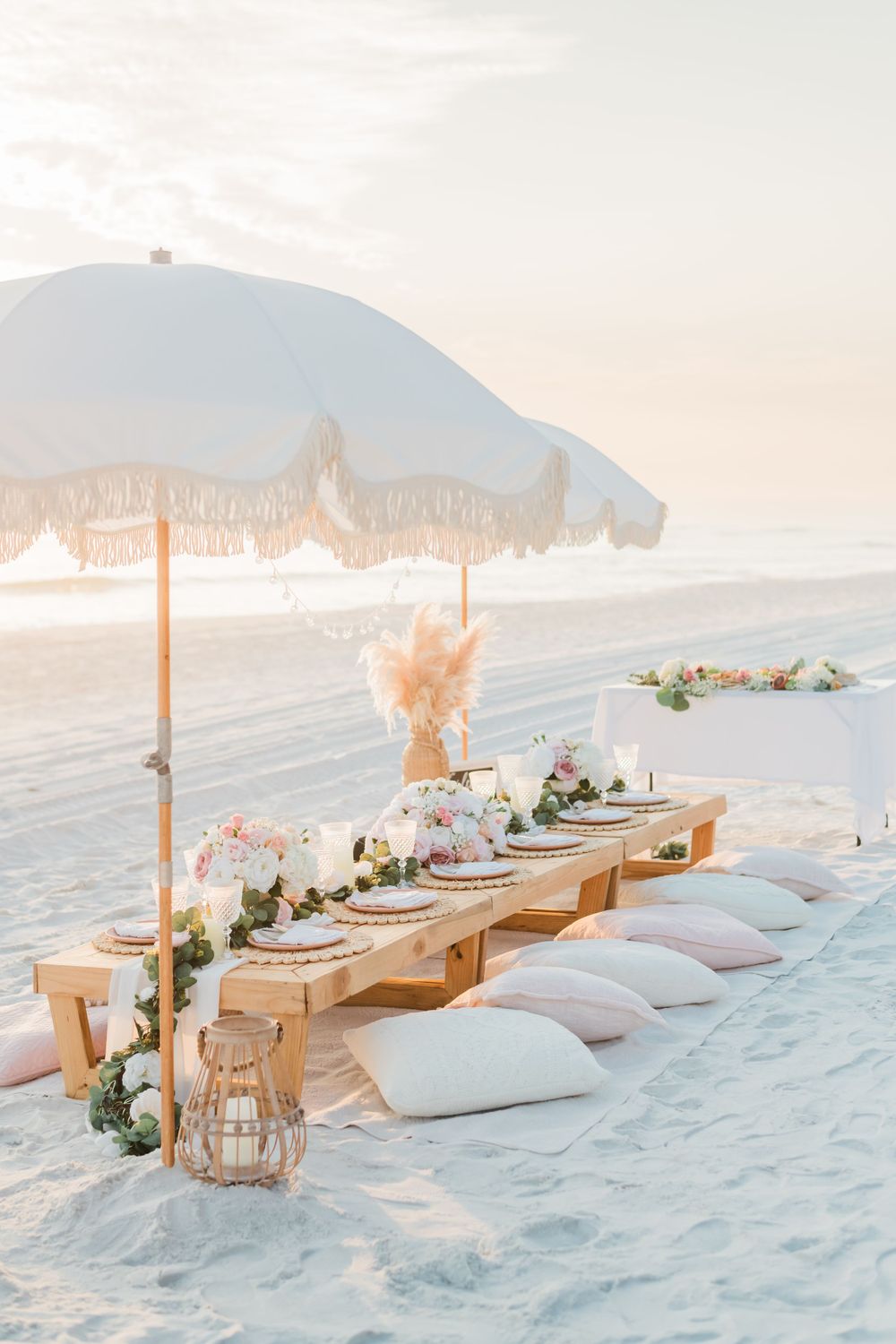 A romantic beach picnic setup with tufted floor cushions, wooden table, parasol and floral arrangements on white sand.