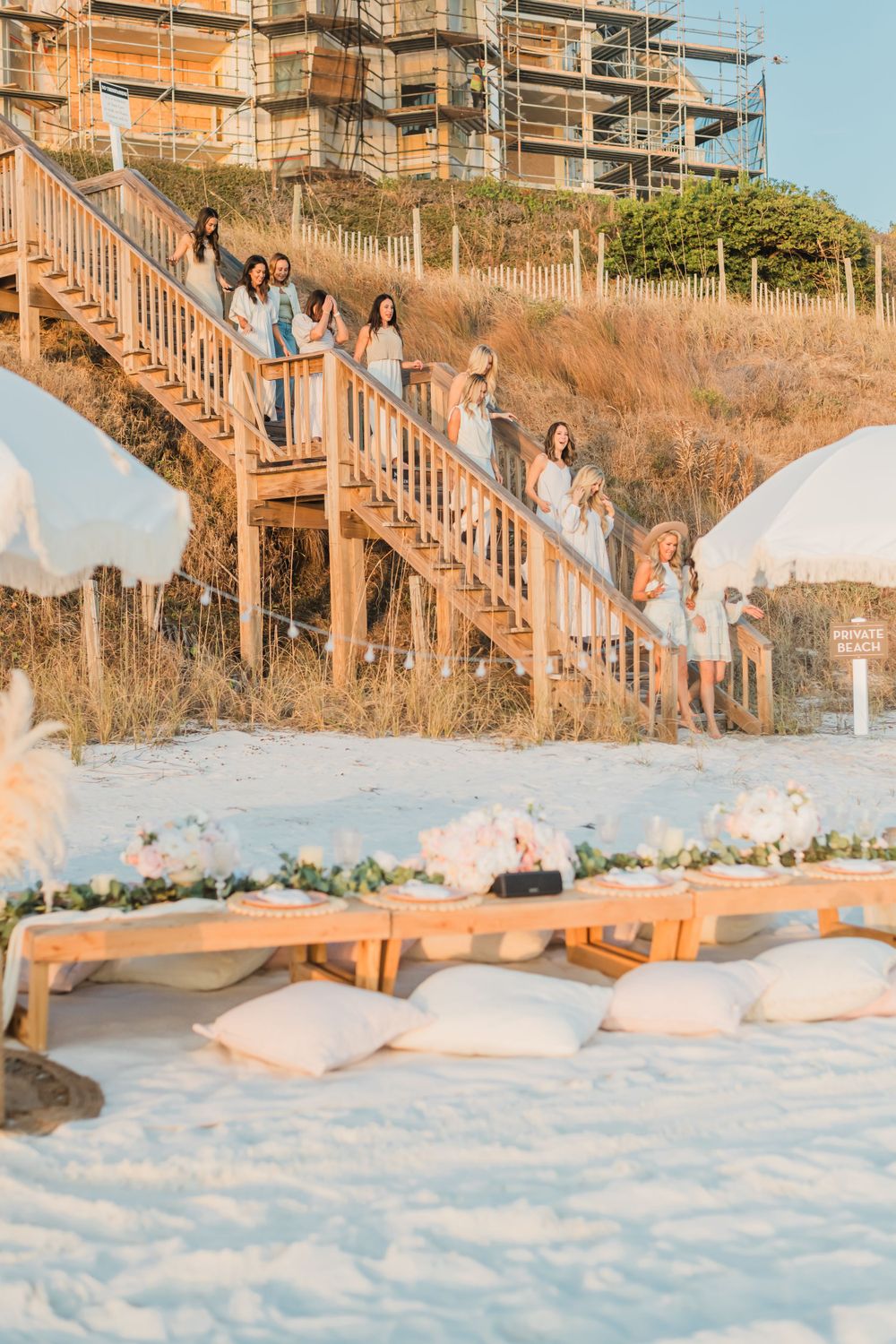 Group stands on wooden beach staircase at sunset with pillows and lounge setup visible on white sand below.