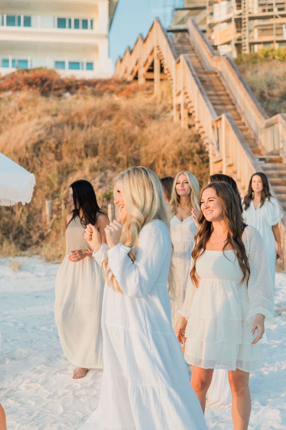 Group wearing white dresses walking along sandy beach at sunset with wooden stairs in background.