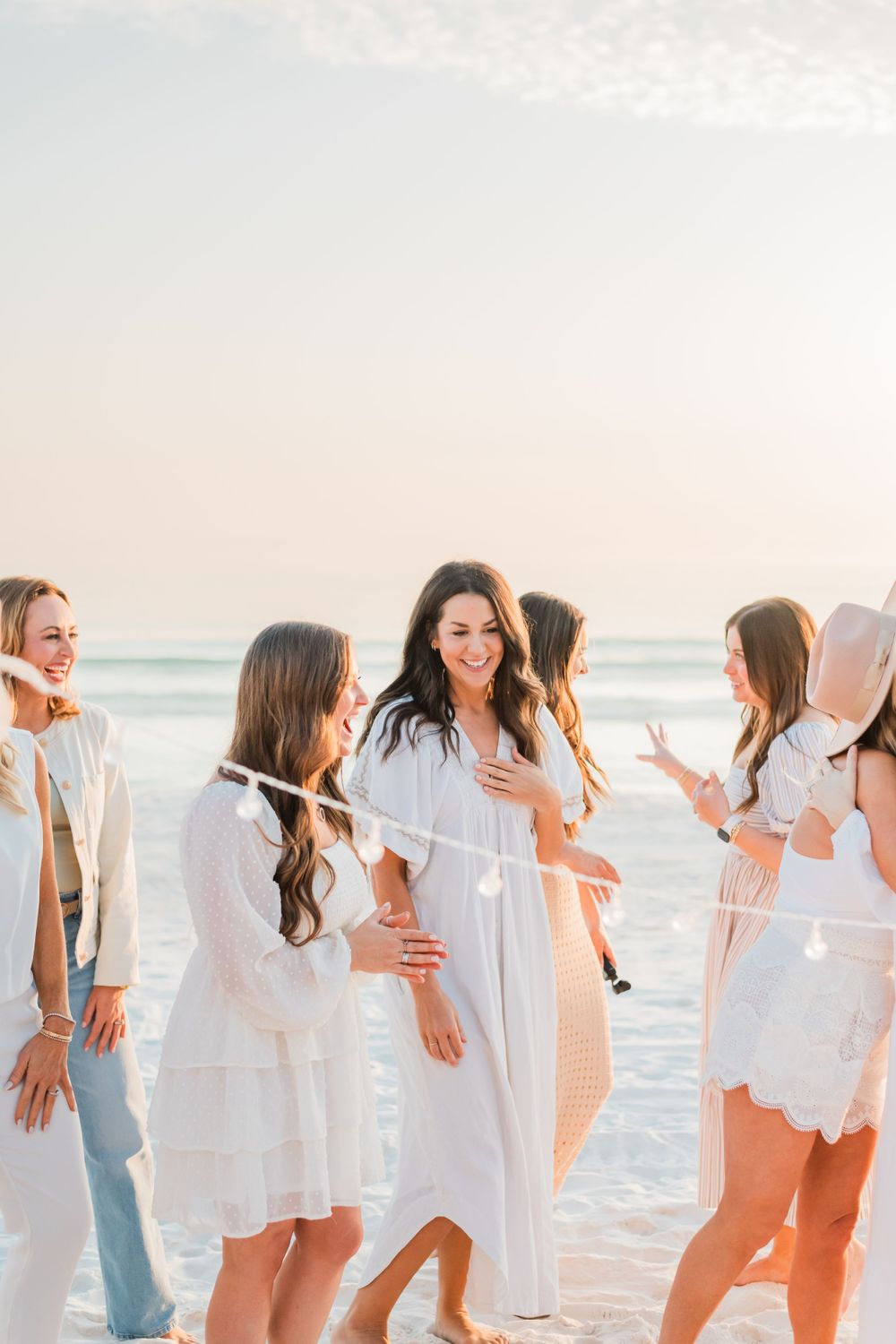 Group of friends in white dresses and casual wear laughing together on a sunny beach at sunset.