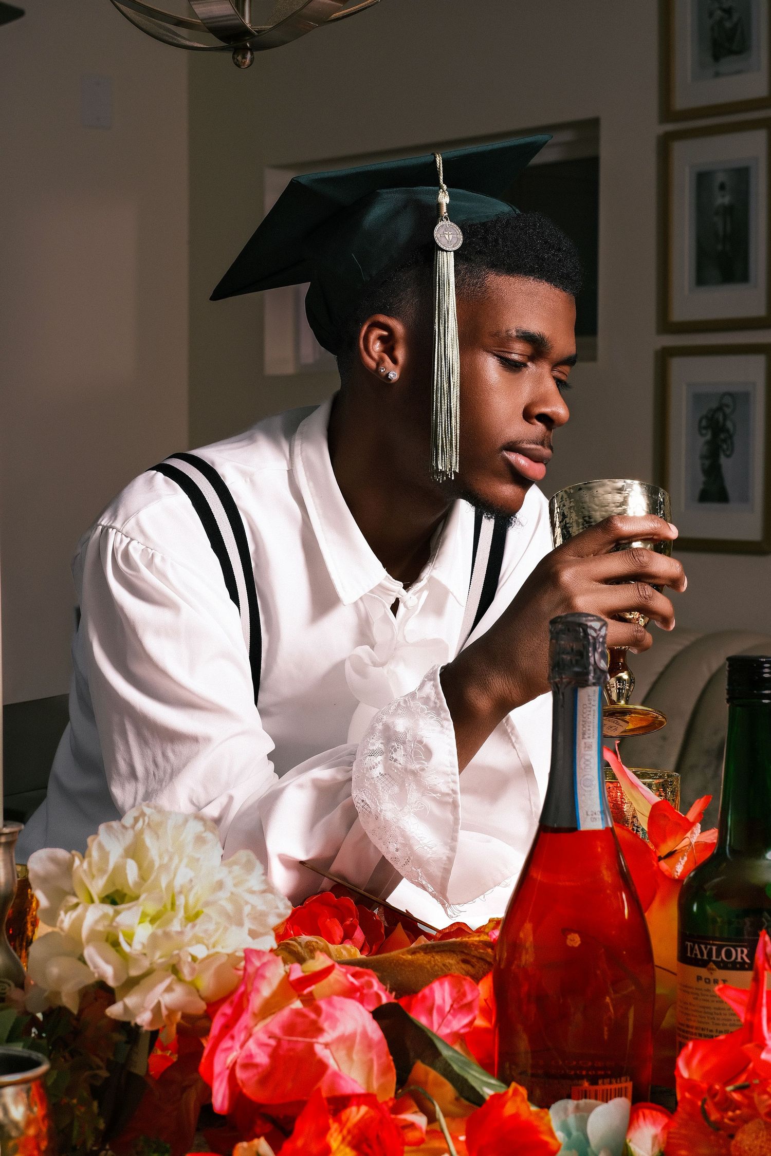 Graduate in cap and gown reflects at table with champagne bottle and red and yellow flowers.