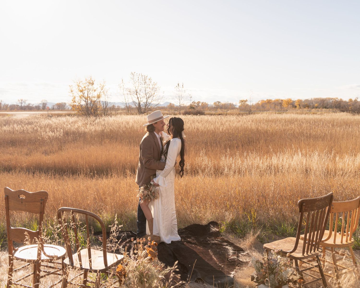 Couple standing among vintage wooden chairs in tall autumn prairie grass at sunset.