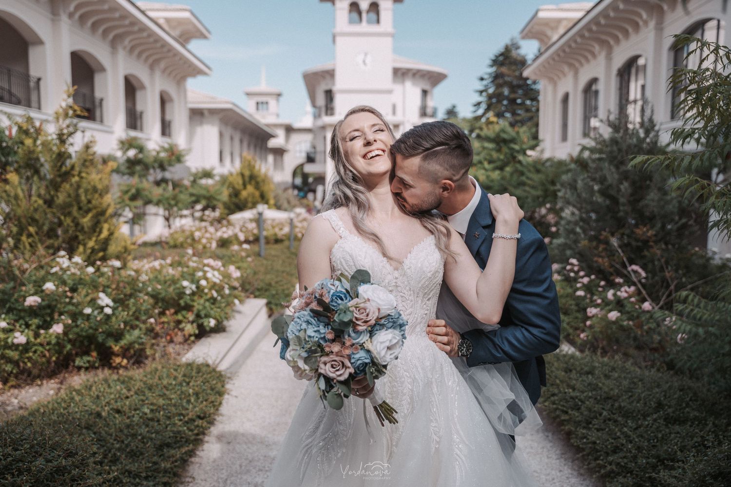 Joyful newlyweds share intimate moment in garden courtyard of white Mediterranean-style building.
