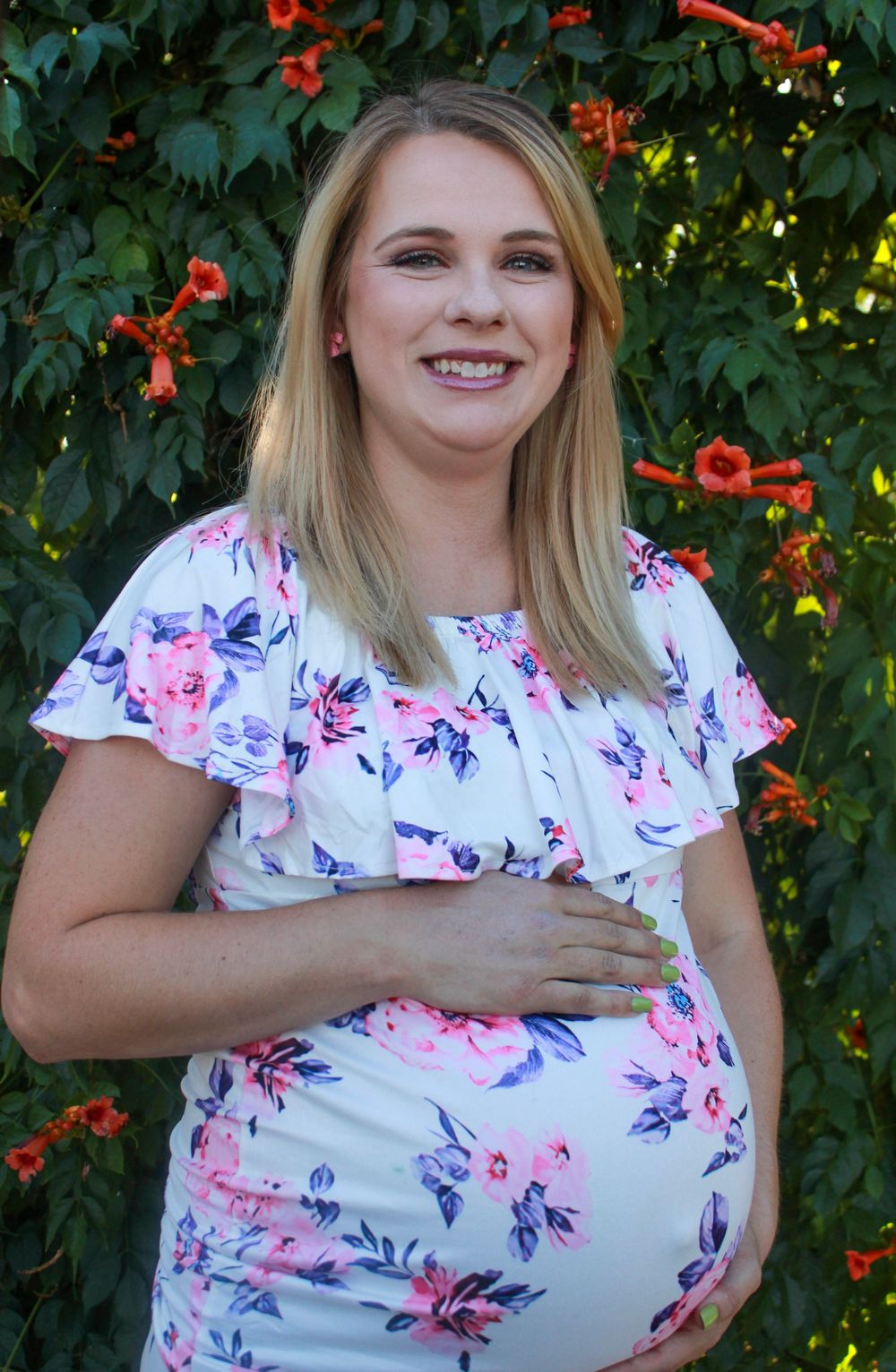 Smiling pregnant woman in floral white dress posing outdoors with red flowers and green foliage background.