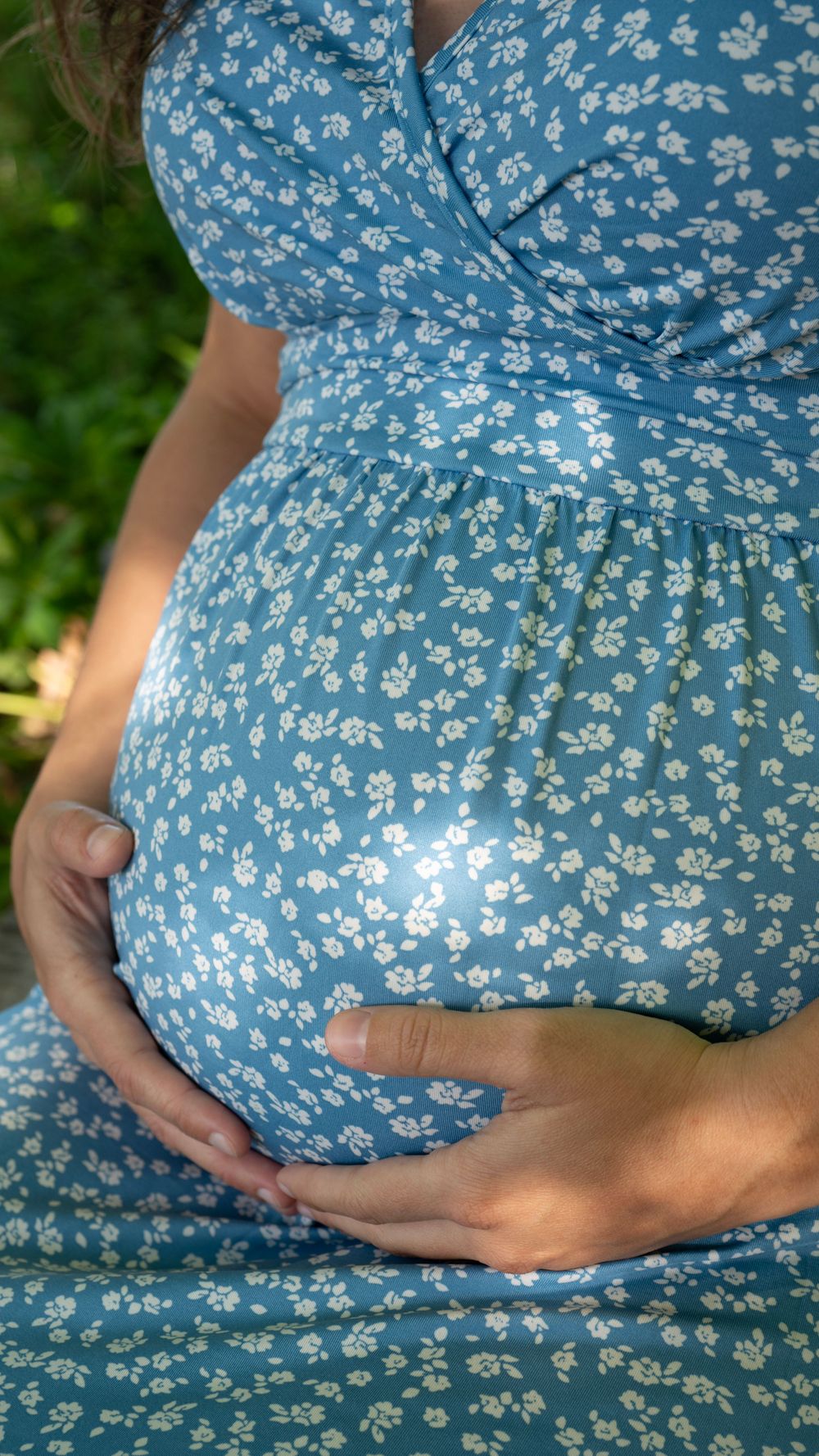 Pregnant woman in blue floral dress gently cradling her baby bump outdoors in natural light.